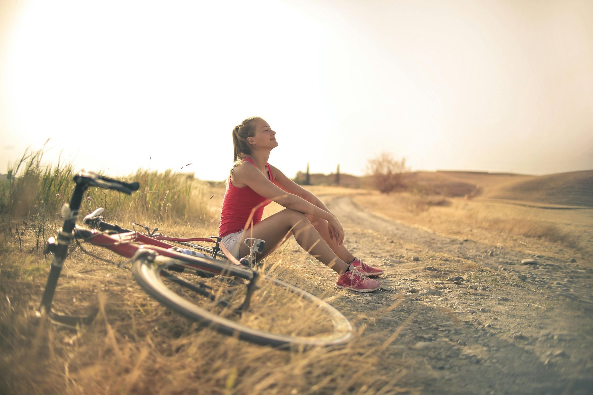 Uma mulher de regata vermelha e shorts brancos descansa ao lado de uma bicicleta em uma estrada de terra, olhando para o céu.