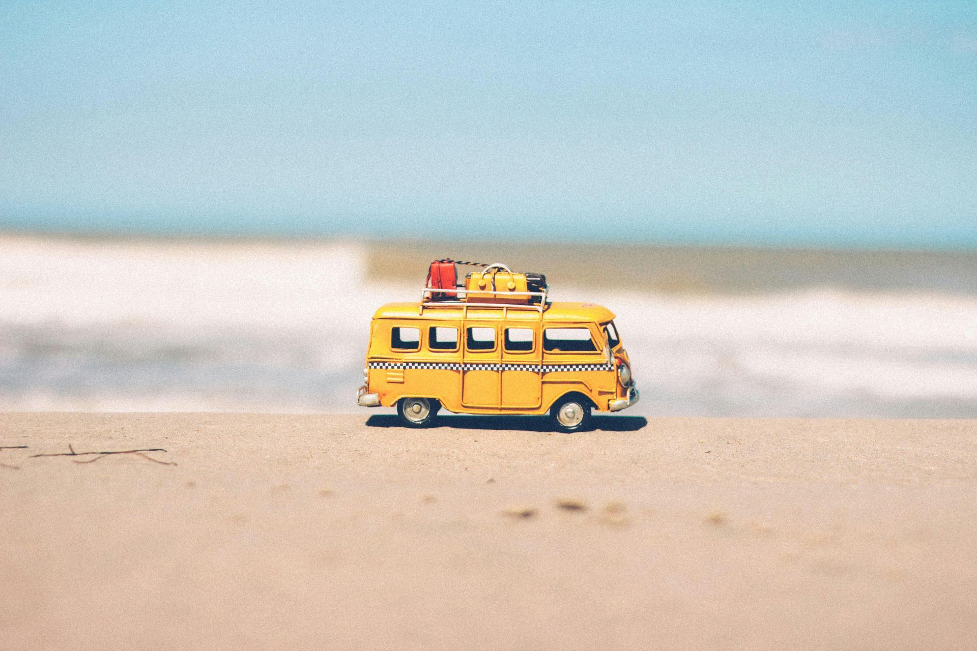 Yellow toy bus with luggage on a beach, waves in background, clear blue sky.