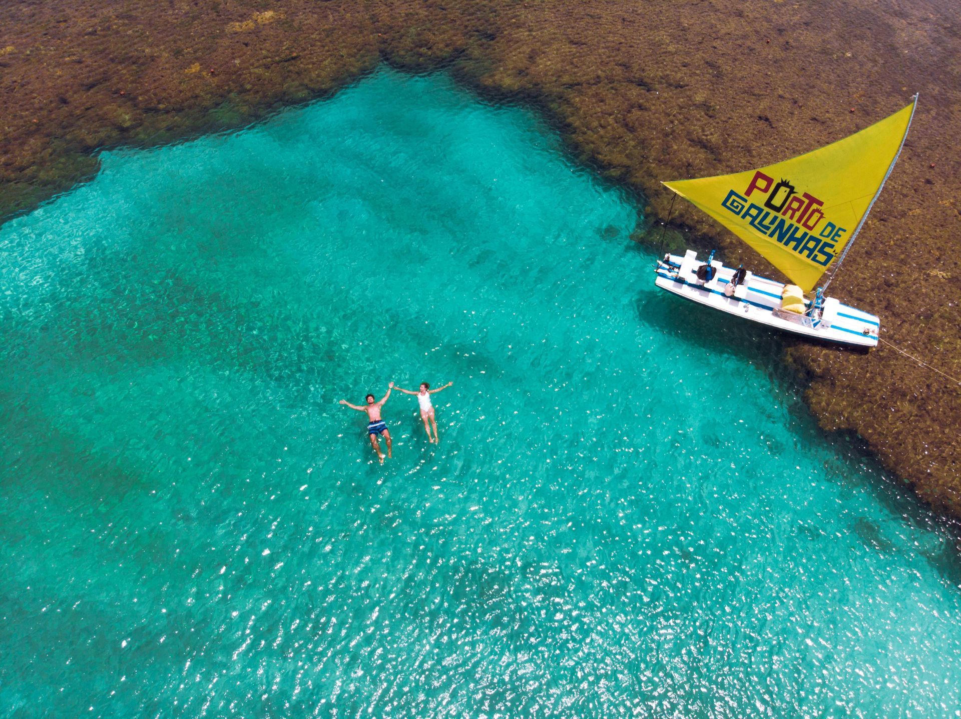 Two people swim in turquoise water near a yellow-sailed boat, Porto de Galinhas, set against a rocky reef.