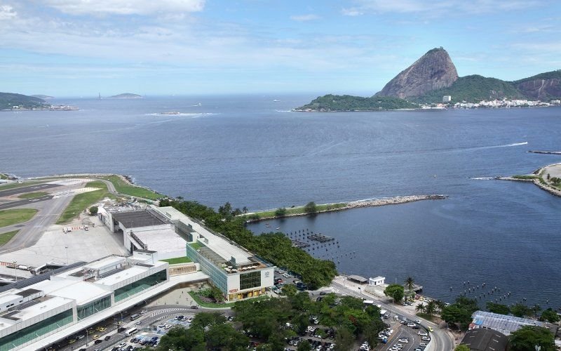 Vista aérea do Rio de Janeiro com o Pão de Açúcar ao fundo, aeroporto, baía e paisagem urbana sob um céu azul.