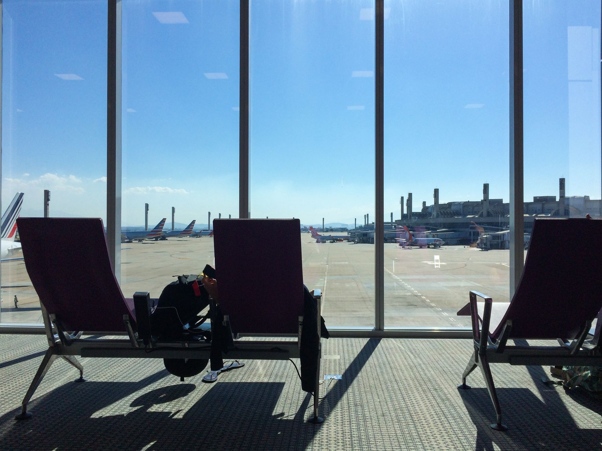 Área de assentos do aeroporto com vista para a pista. Pessoa na cadeira central, céu azul, aviões visíveis.