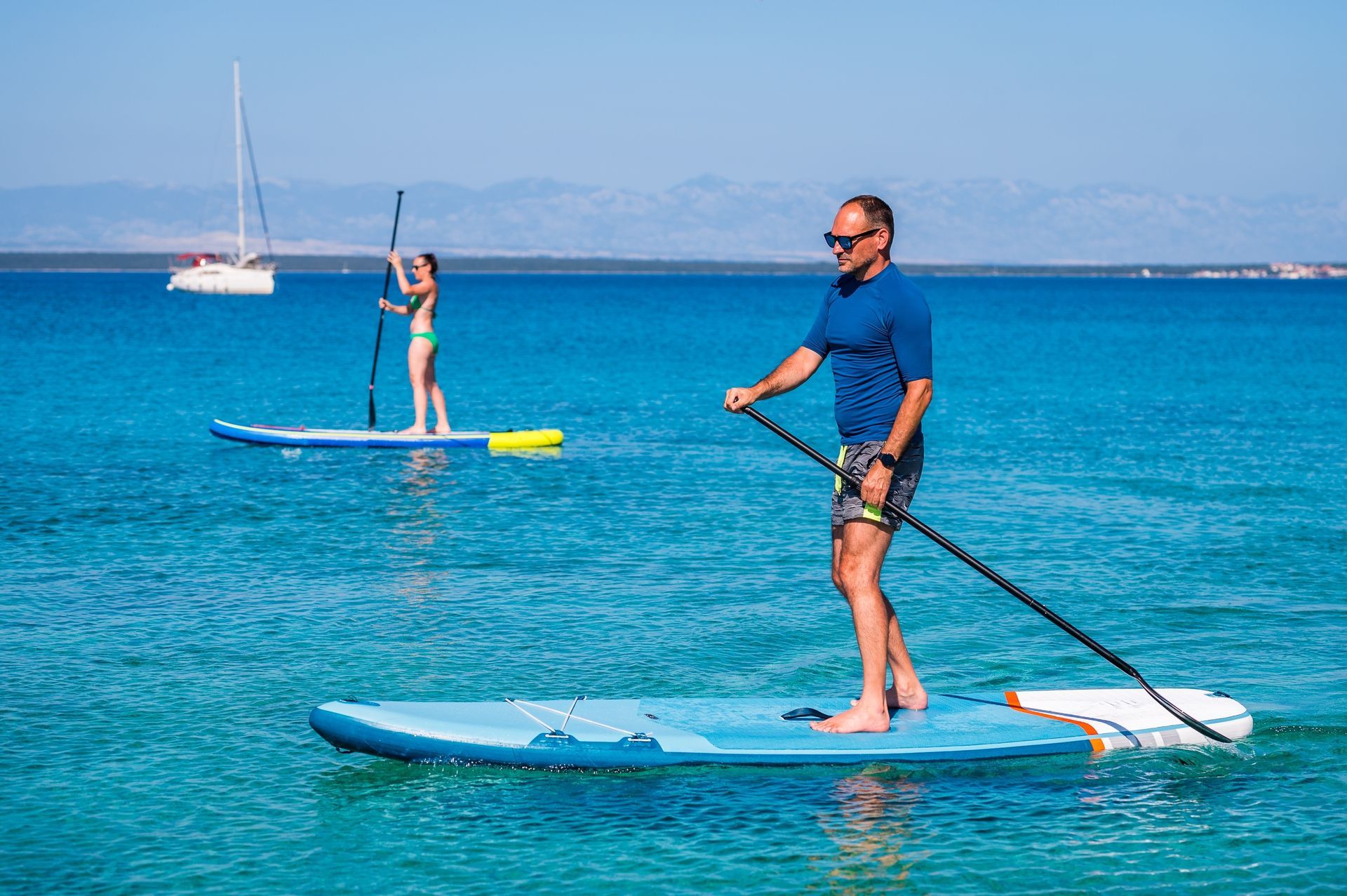 Man stand-up paddleboards in clear blue water, another person further back. Boat in background, mountains on the horizon.