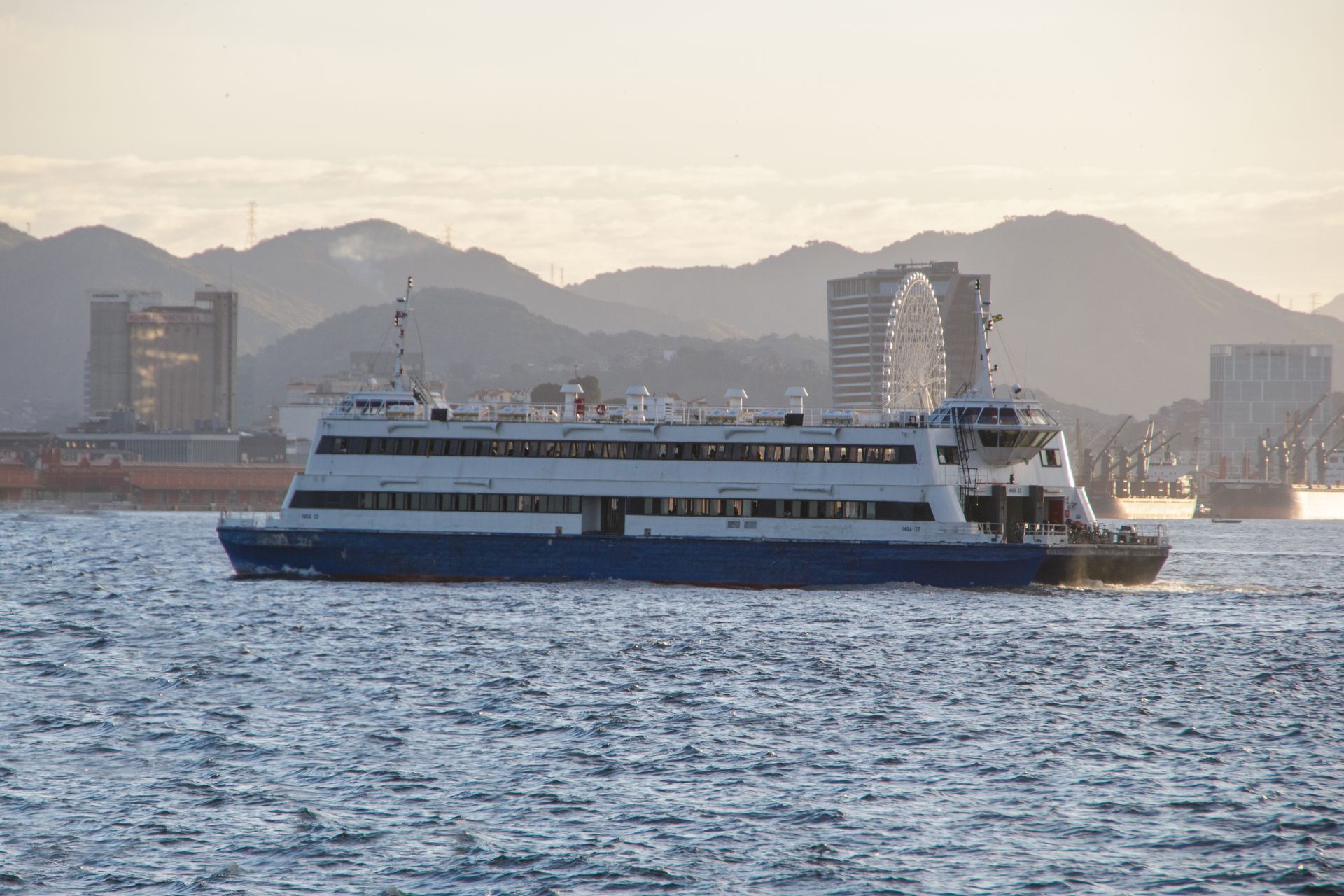 Ferry boat on water, with city buildings and mountains in the background at sunset.