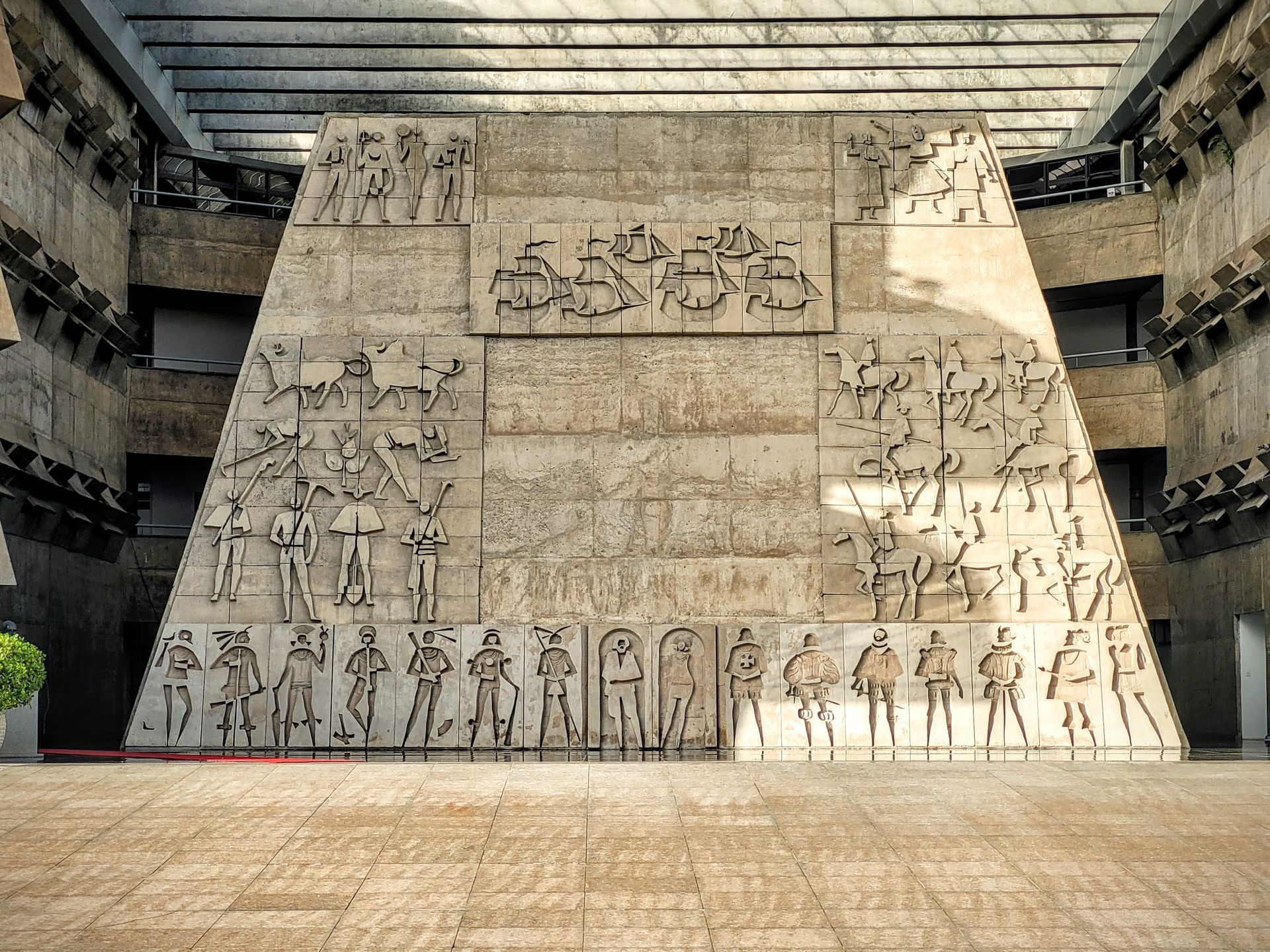 Large stone mural with carved figures in a modern atrium, viewed from across the room.