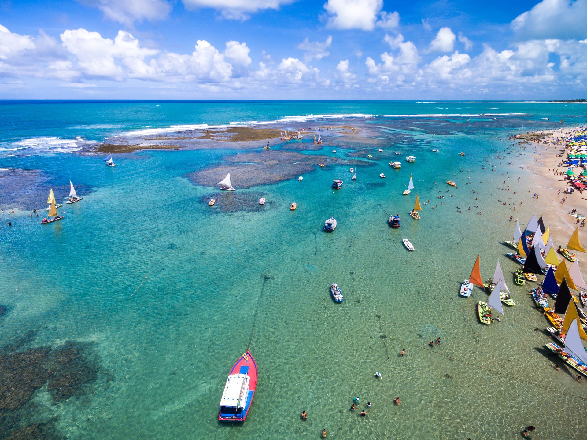 Aerial view of a turquoise beach with boats, swimmers, and a blue sky with clouds.