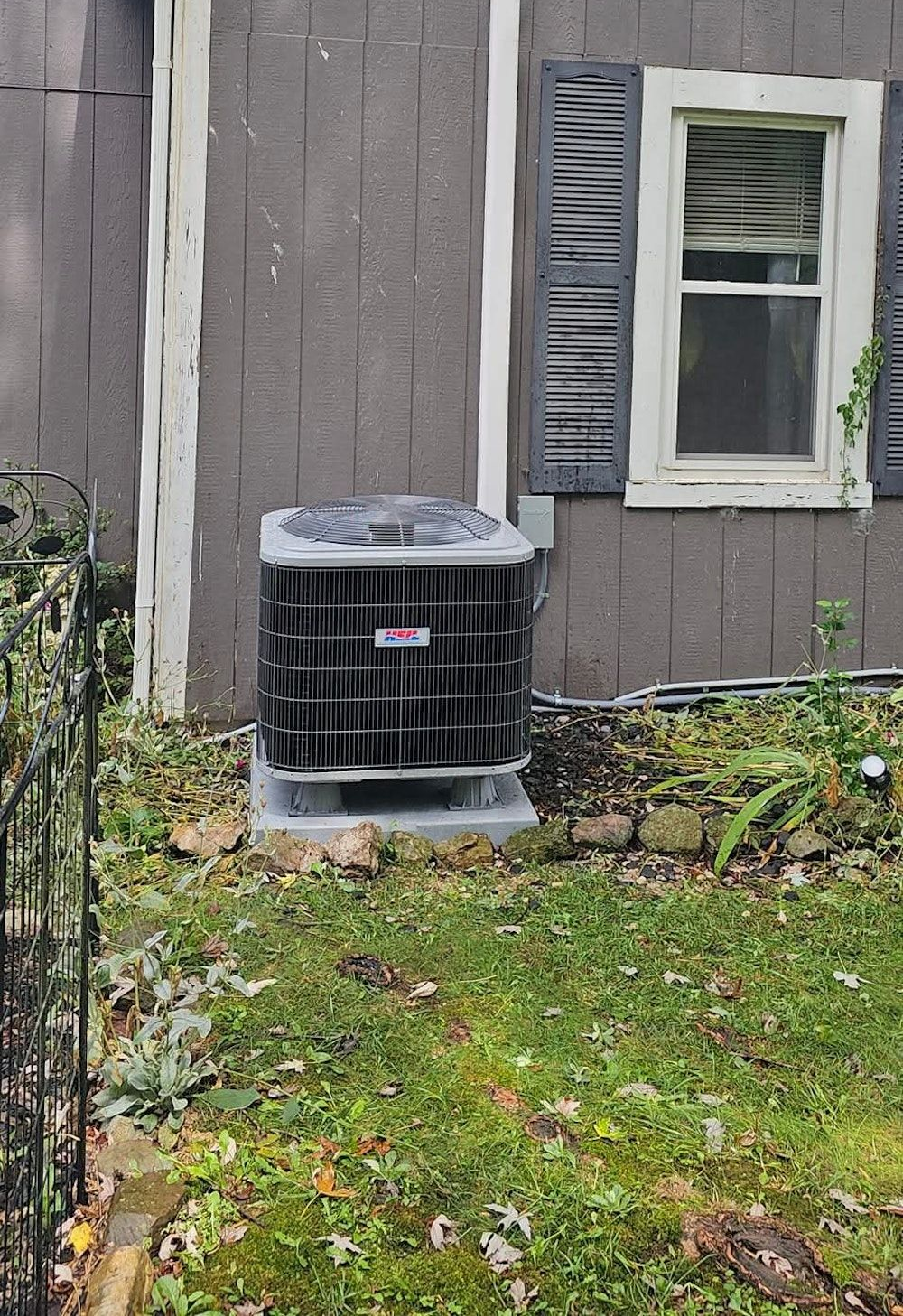 Outdoor AC unit beside a gray house wall with a window and grass in front
