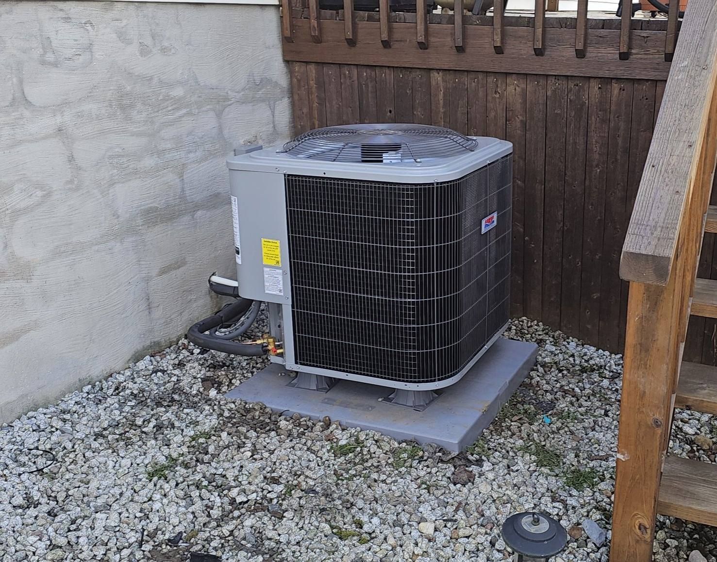 Outdoor HVAC unit beside a stucco wall and wooden fence on a gravel pad