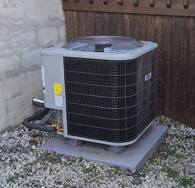 Outdoor central air conditioning unit on a concrete pad beside a stone wall and wooden fence.