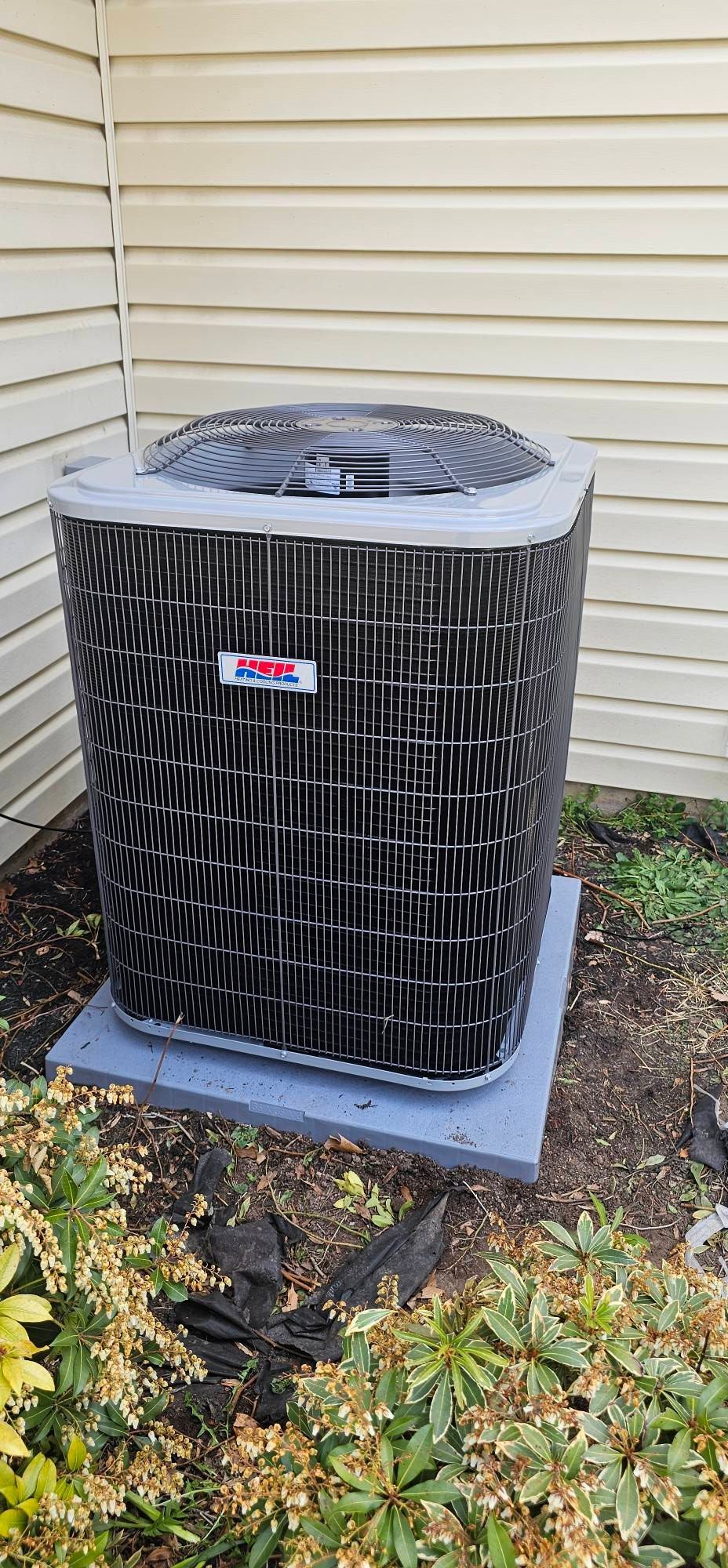 Outdoor air conditioner unit beside beige house siding and plants