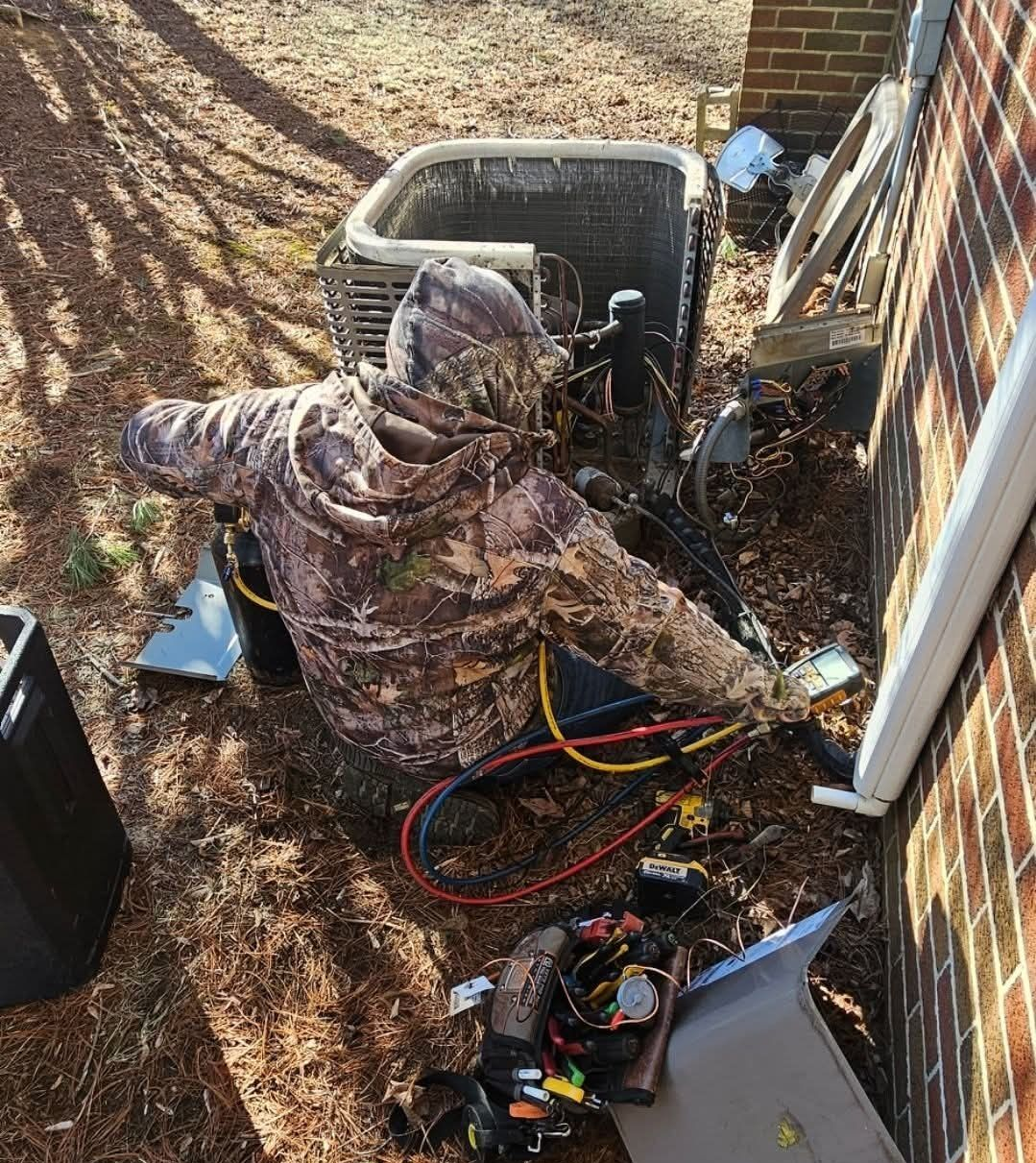 Worker in camouflage jacket clearing debris beside a brick house, with tools and cables on the ground