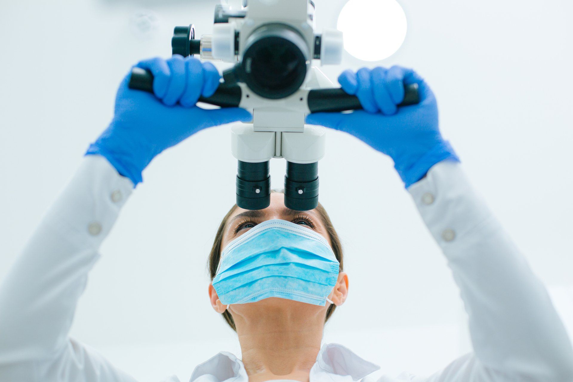 A woman wearing a mask and gloves is looking through a microscope.
