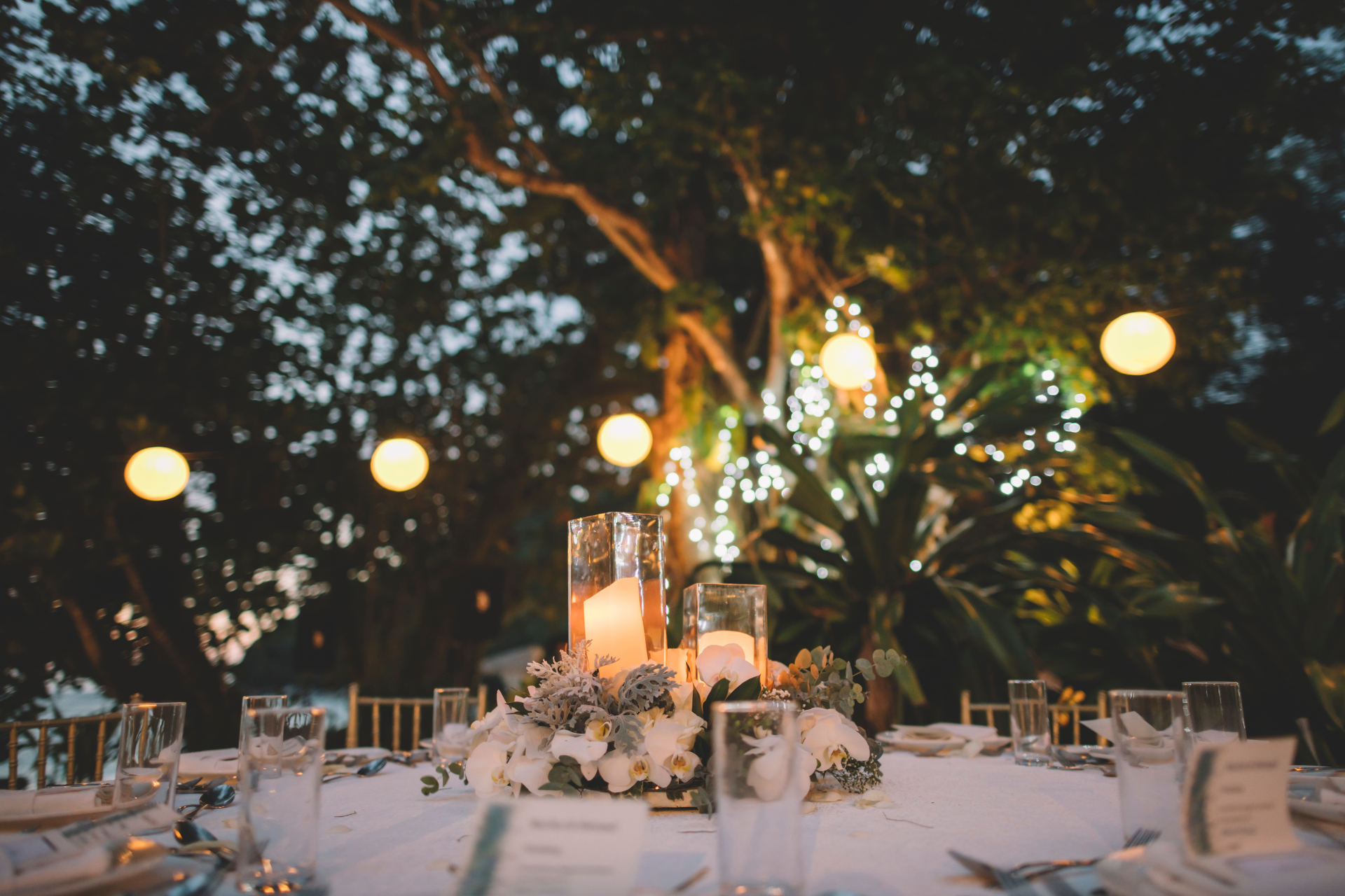 A round, white-clothed table is set outdoors at dusk with a floral centerpiece, glass candles, and hanging paper lanterns.
