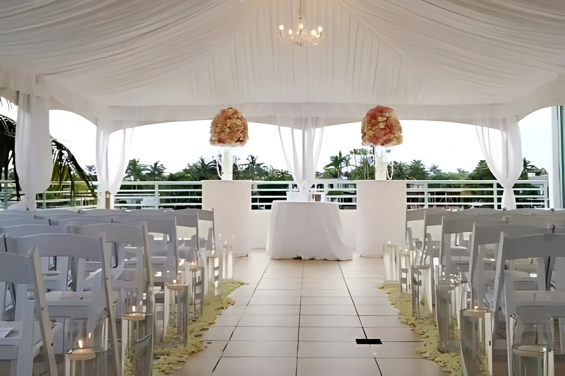 Wedding ceremony setup with white chairs, floral arrangements on pillars, and a draped tent overlooking a tropical view.
