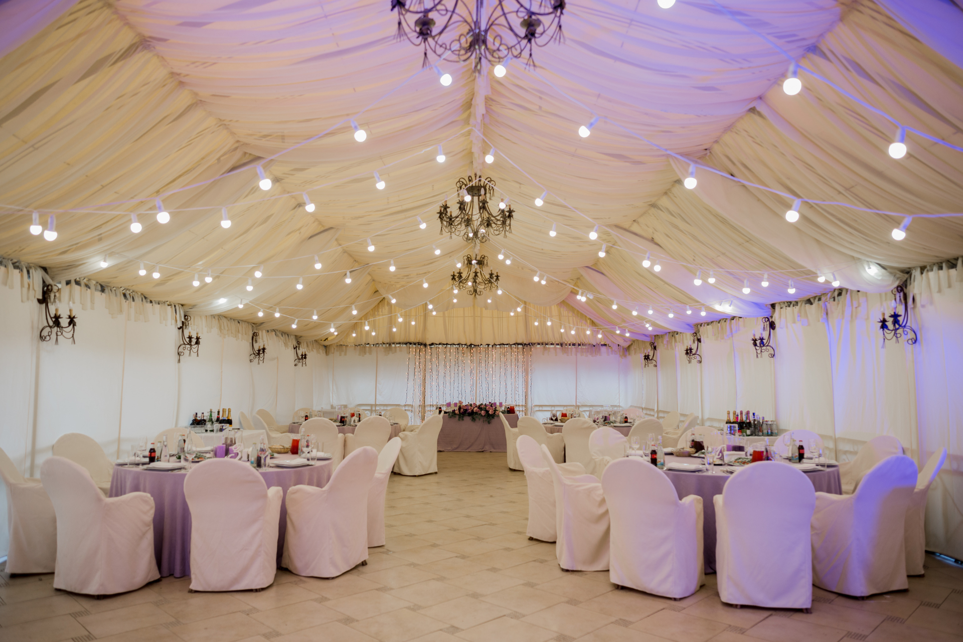 A draped event tent interior with round tables, white covered chairs, and chandeliers illuminated by fairy lights.