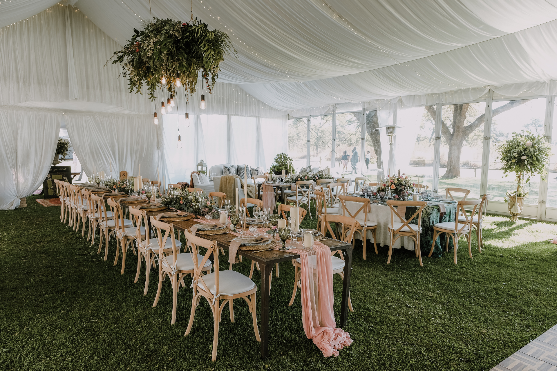 An outdoor tent set up for a wedding reception with long tables, wooden chairs, and a hanging greenery floral centerpiece.