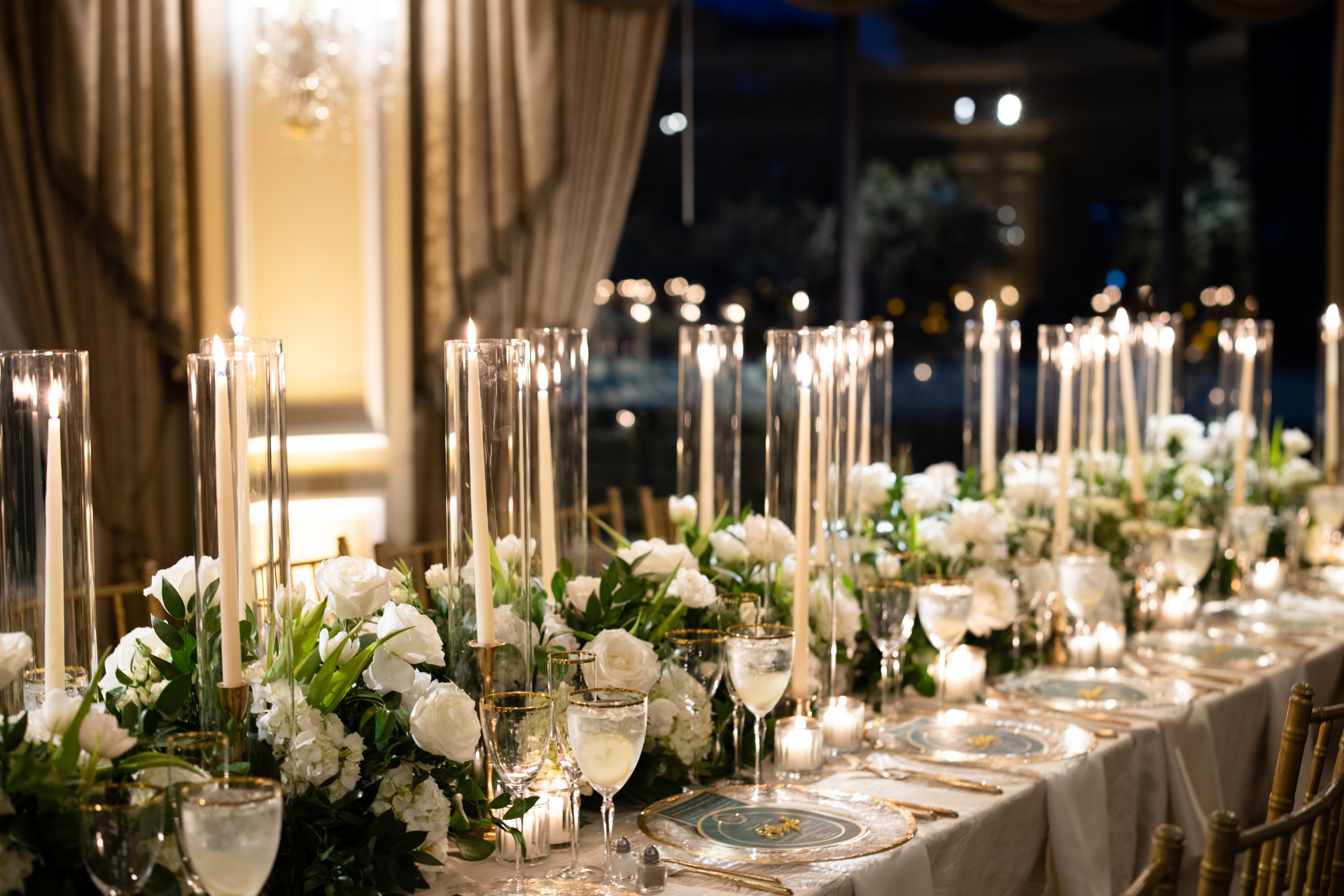 A long banquet table set with white floral arrangements, tall candles in glass cylinders, and elegant place settings.