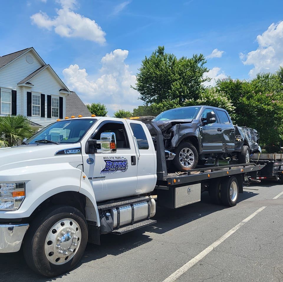 Tow truck hauling a damaged black SUV on a flatbed beside a suburban house under a blue sky