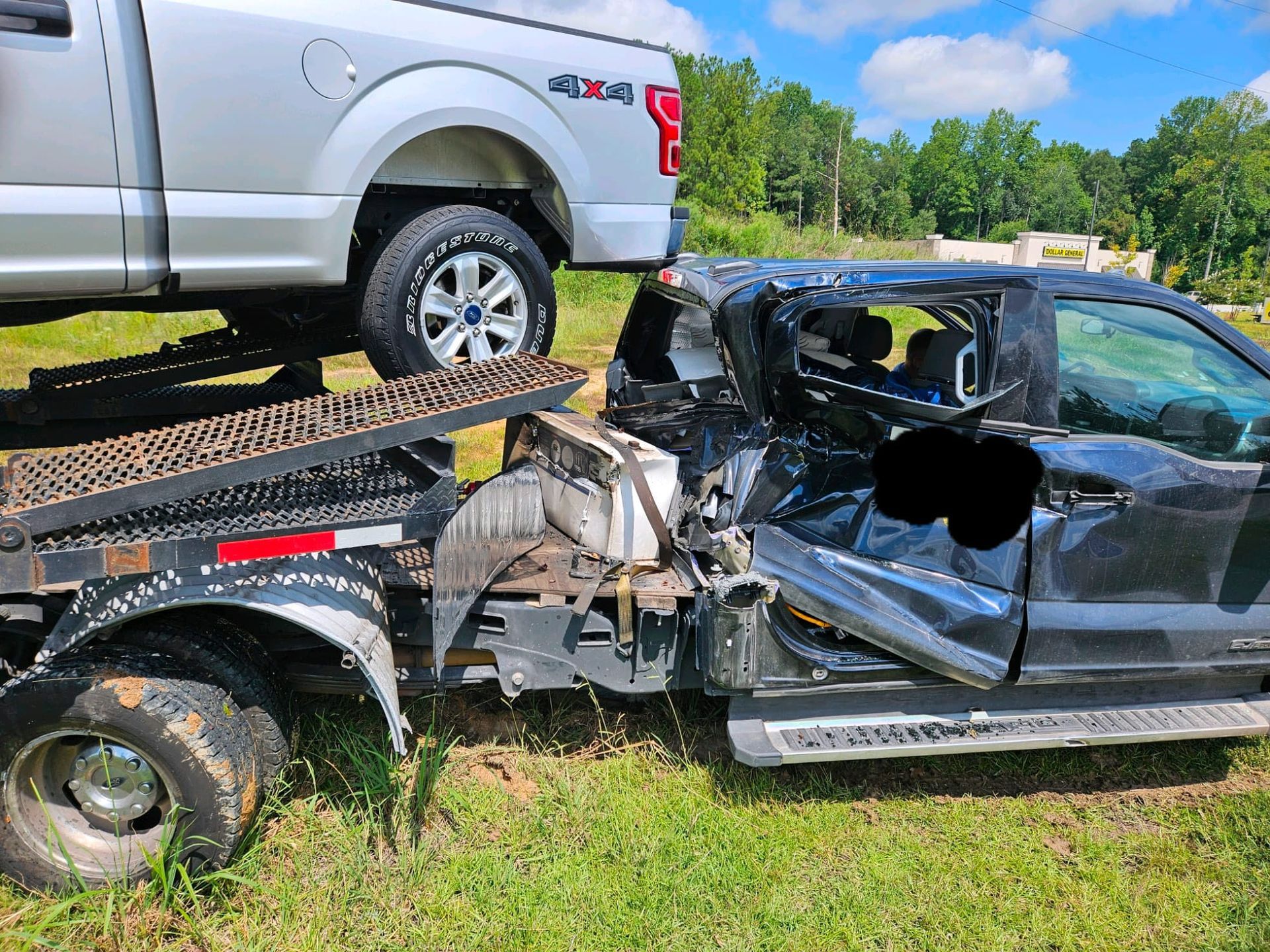 Wrecked black car pinned under a silver pickup on a tow trailer in a grassy field