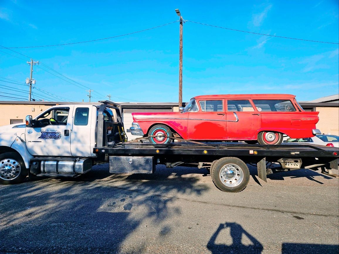 Tow truck carrying a white pickup and red vintage wagon on a flatbed in a sunny parking lot