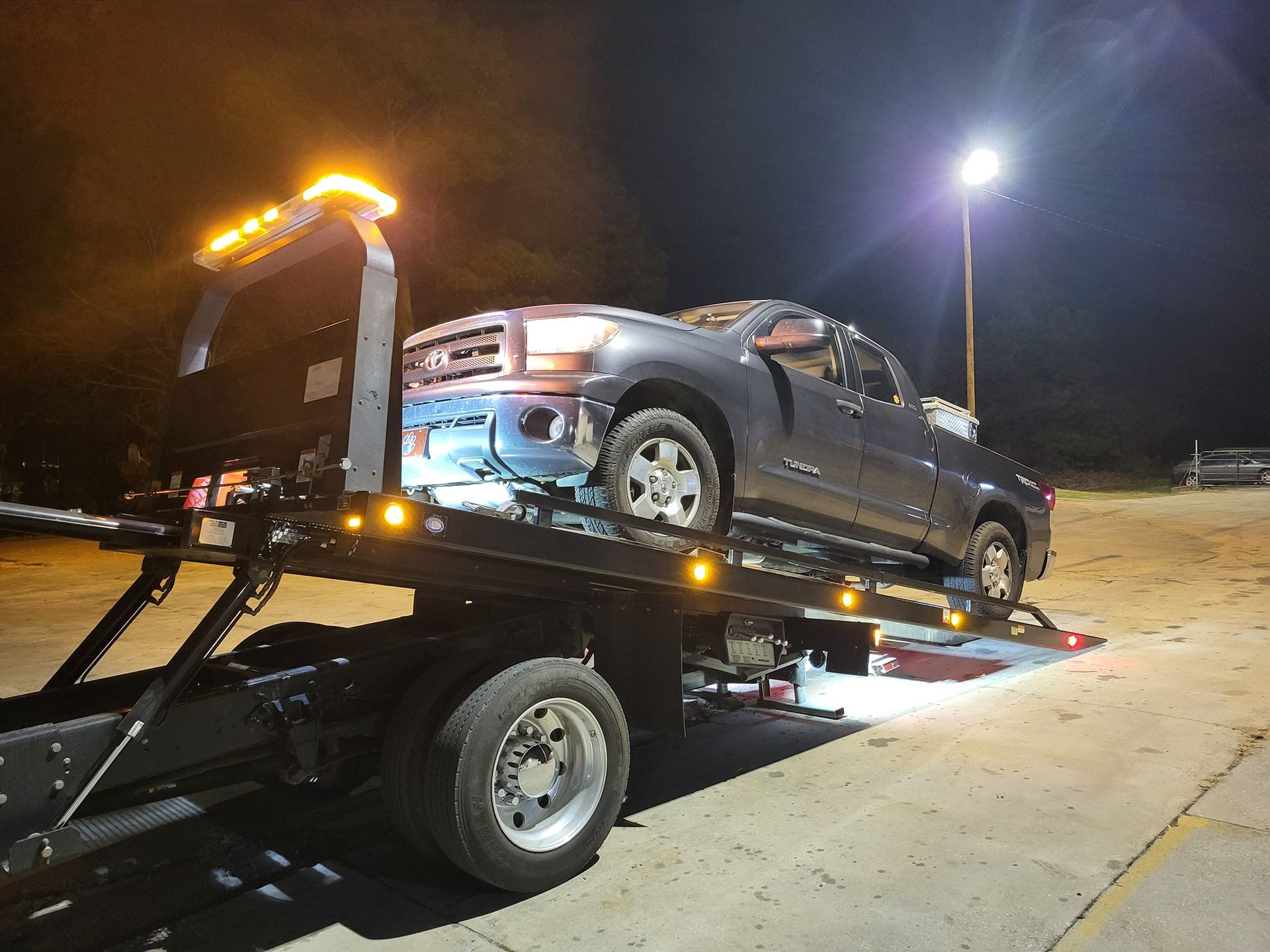 Silver pickup truck on a tow trailer at night, lit by streetlights on a roadside.