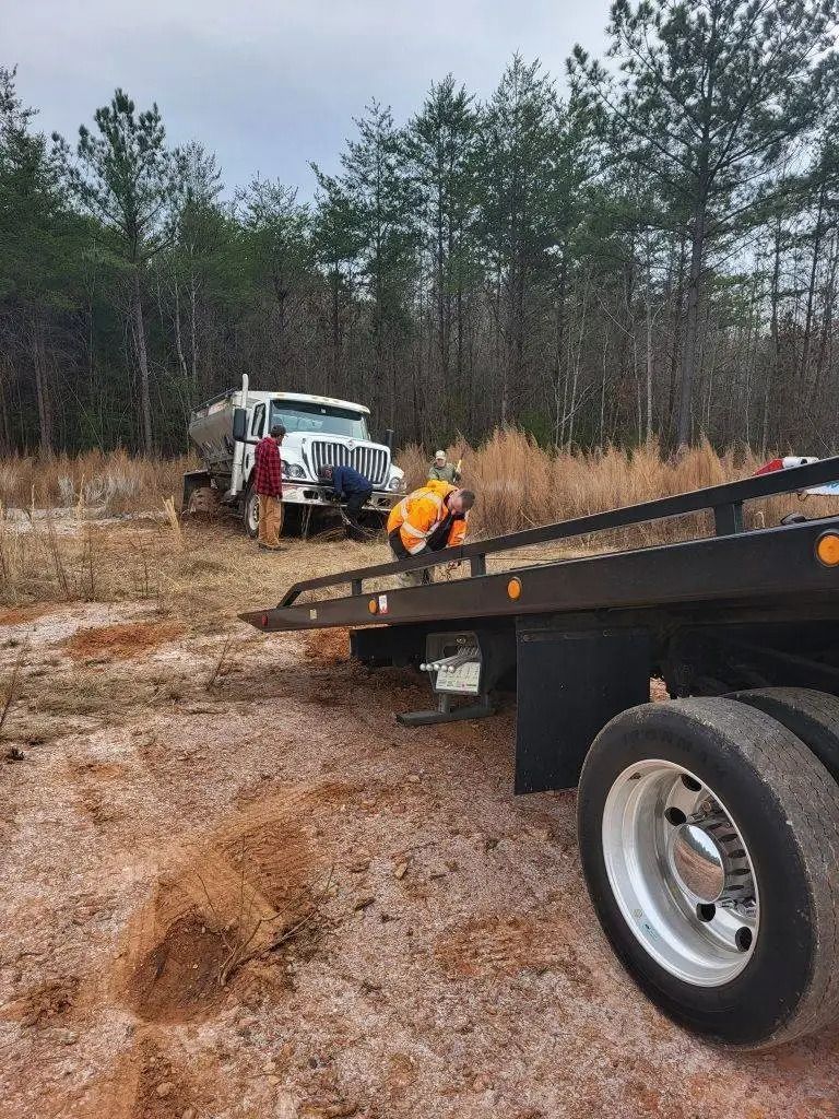 Tow truck and worker in orange vest beside a flatbed trailer on a muddy forest road