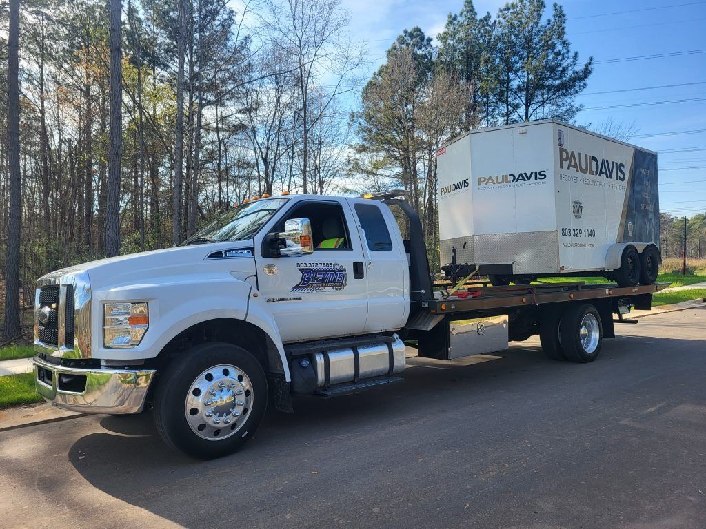 White moving truck with box trailer parked on a road beside trees, with a driver in the cab.