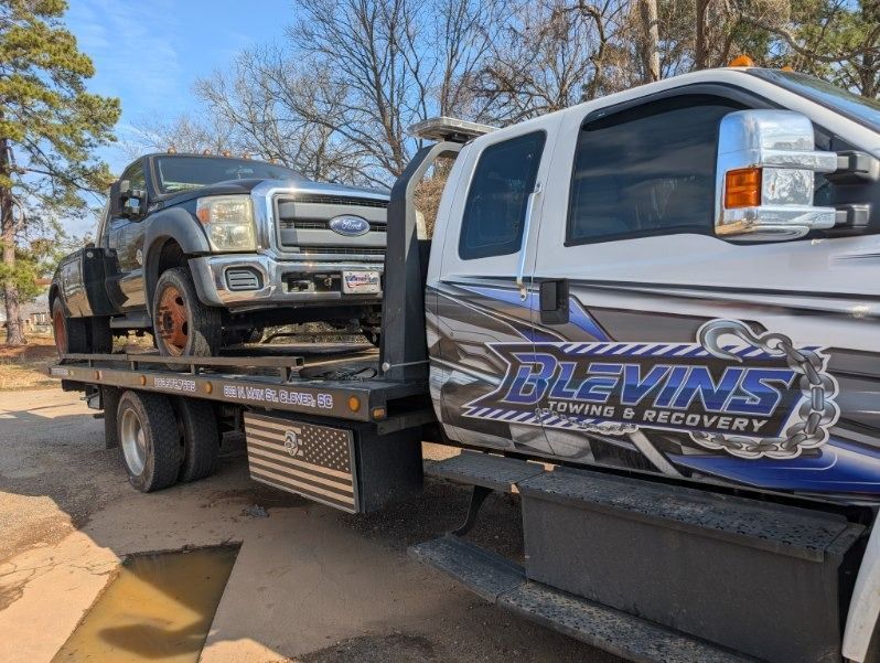 Tow truck hauling a silver SUV on a flatbed in a sunny outdoor lot with Blevins Recovery logo.