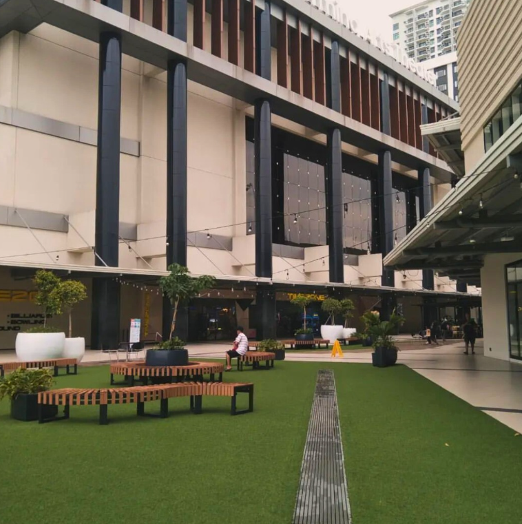Exterior of a modern shopping center, featuring an outdoor seating area with green turf and benches.