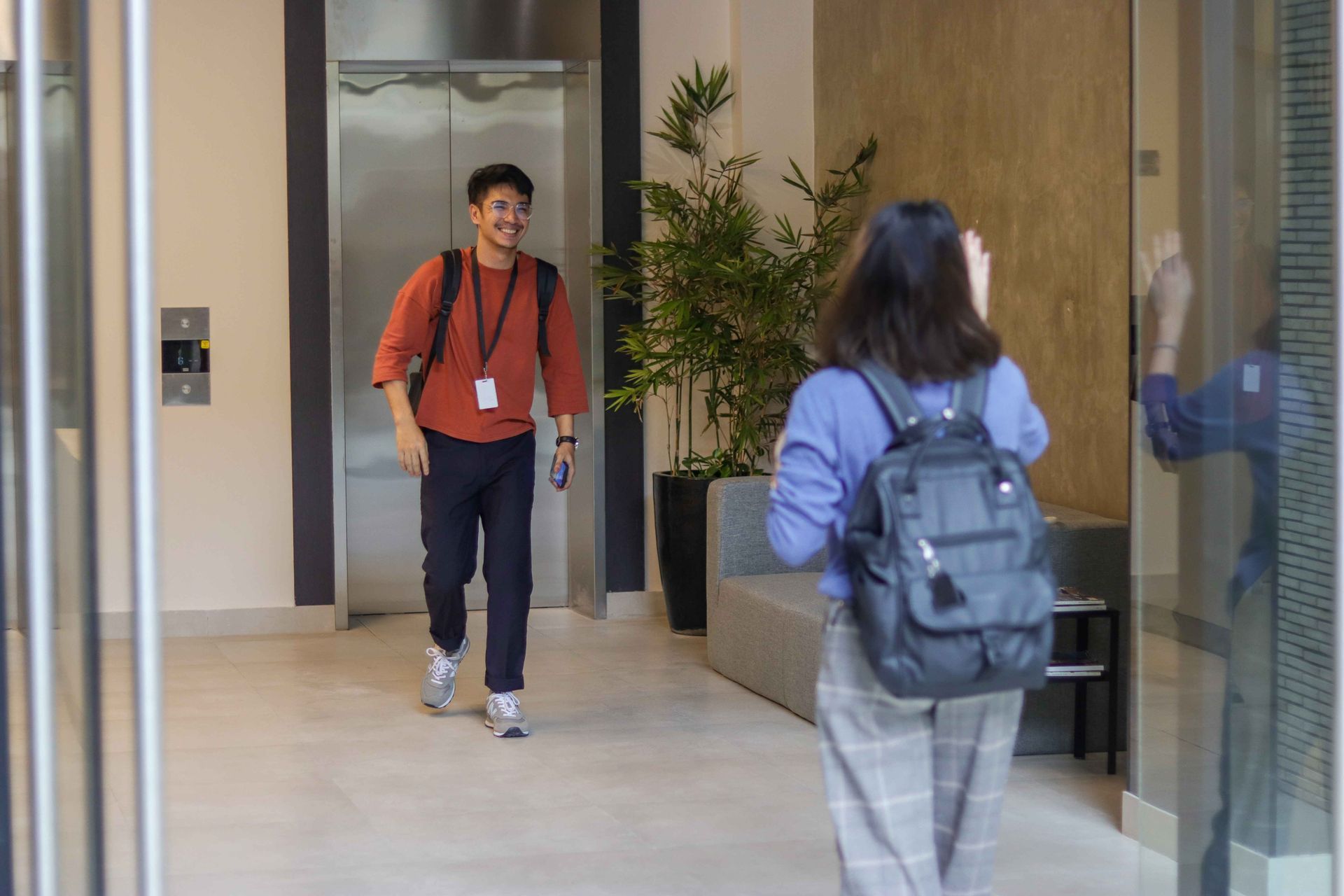 Man approaching woman in lobby; man smiles, woman wears backpack, near elevator and potted plant.