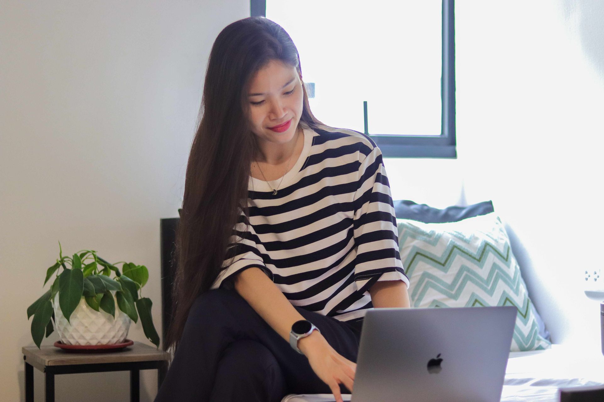 Woman with long hair, working on a laptop in a room, wearing a striped shirt and a smartwatch.