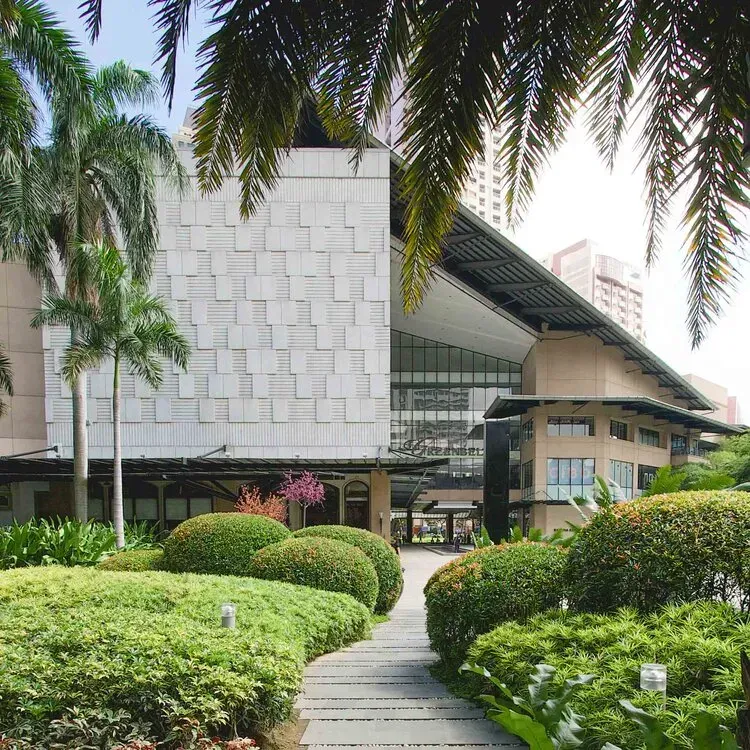 A stone-tiled path winds through manicured shrubs toward a modern building with a textured facade and an overhanging roof.