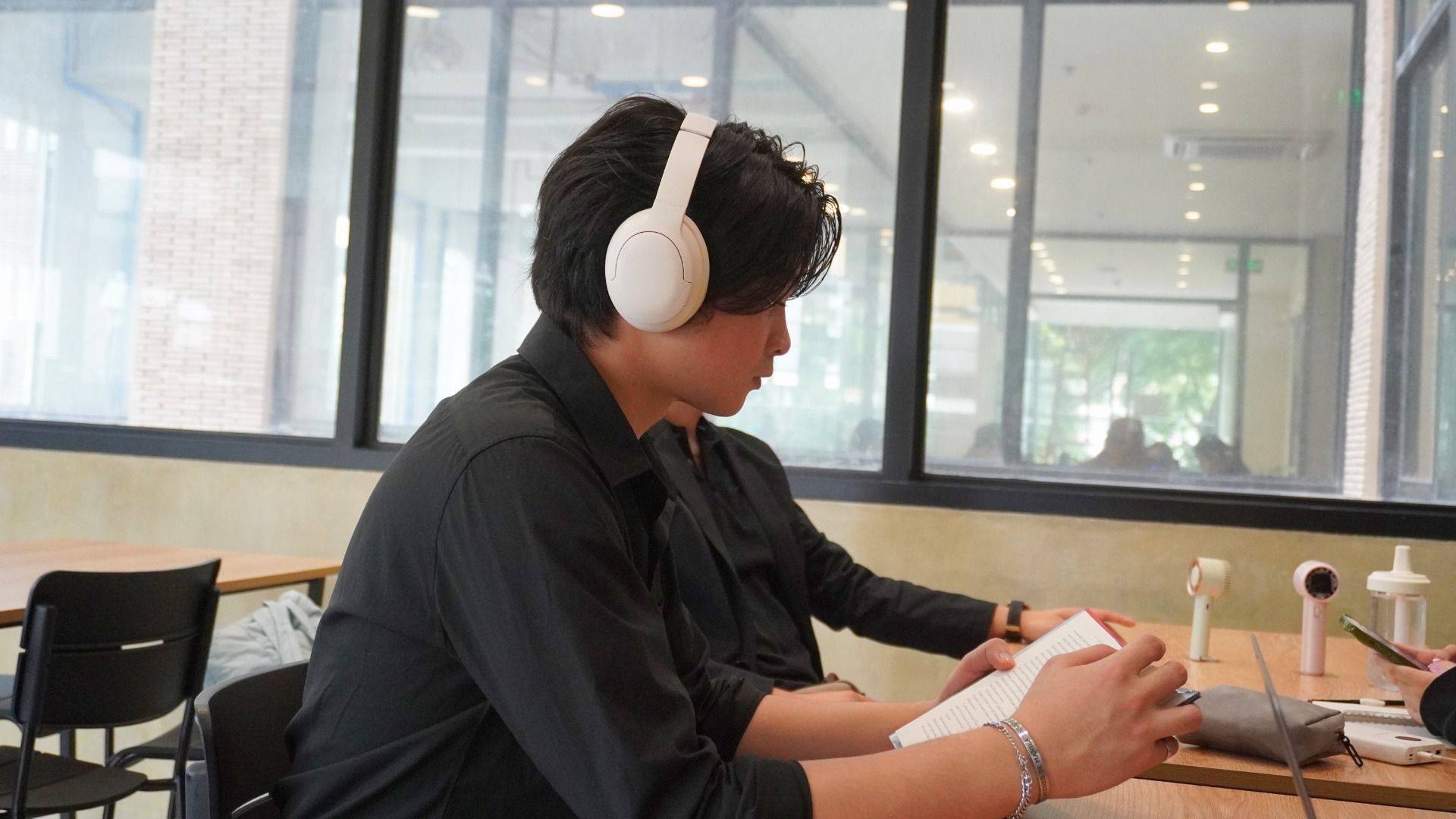 Young person with white headphones reading a book at a table near a window.