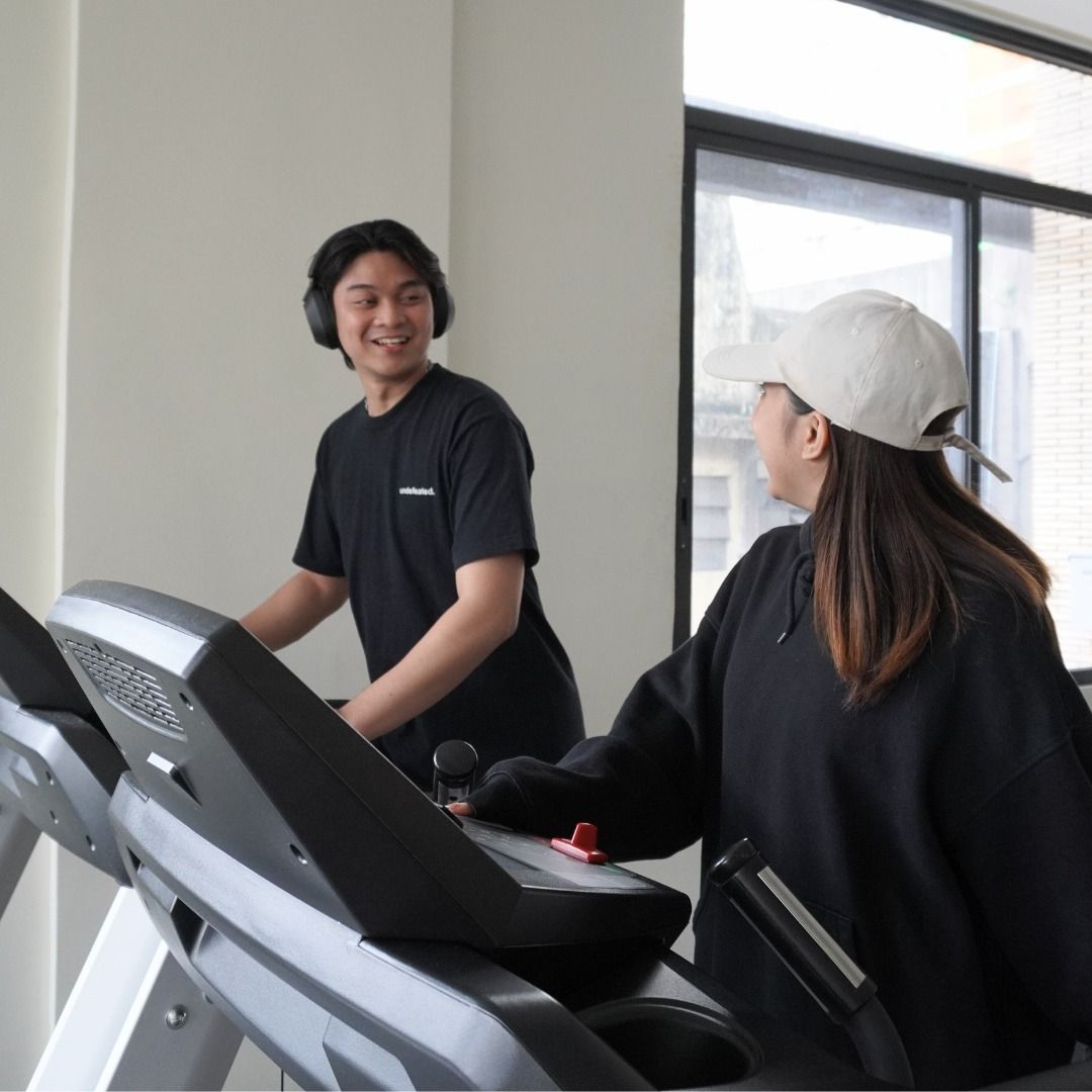 Two people on treadmills in a gym. A man in black shirt and headphones smiles. A woman in a cap faces him.
