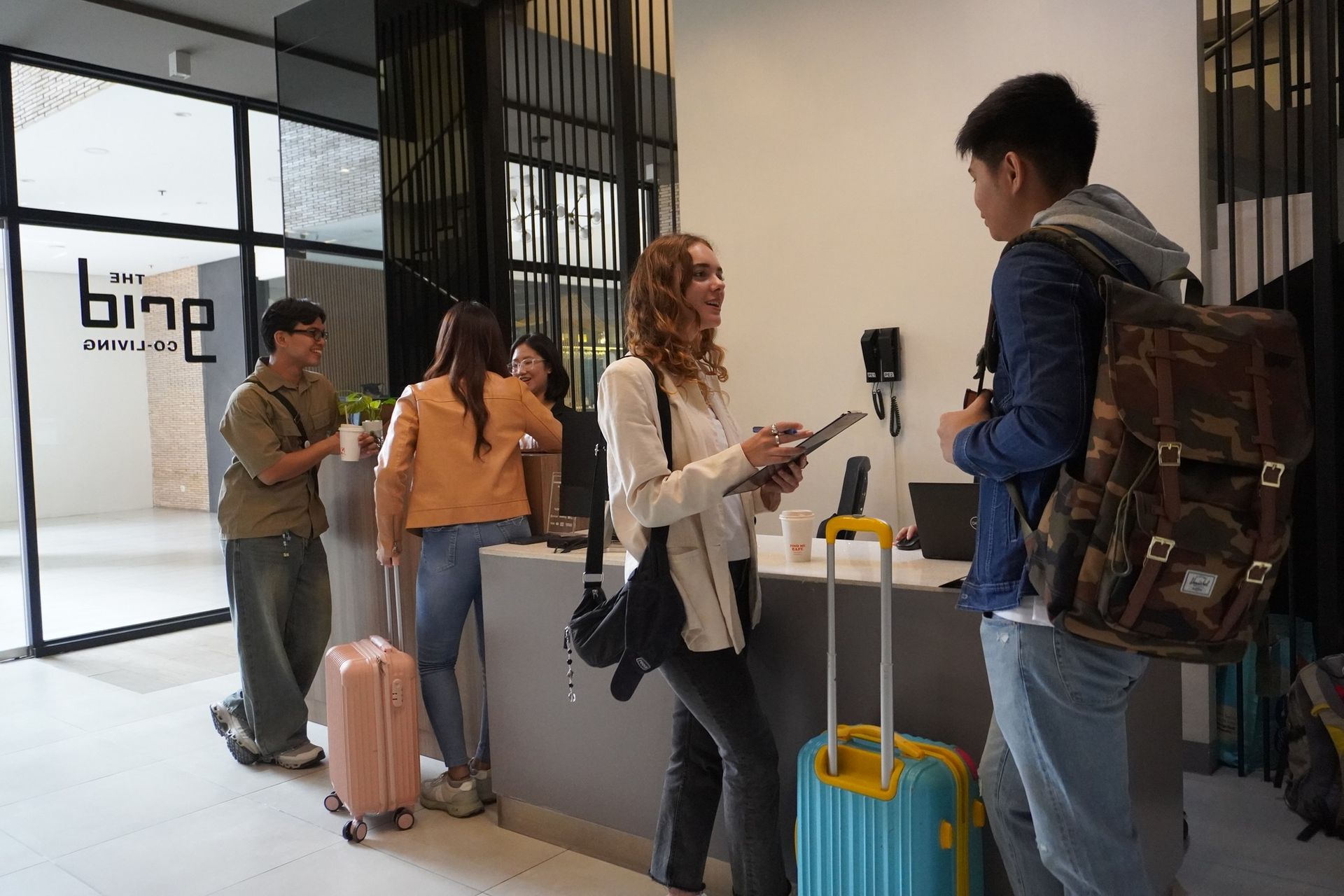 People checking into a hotel, luggage visible. A woman holds a tablet, interacting with a guest. Modern reception area.