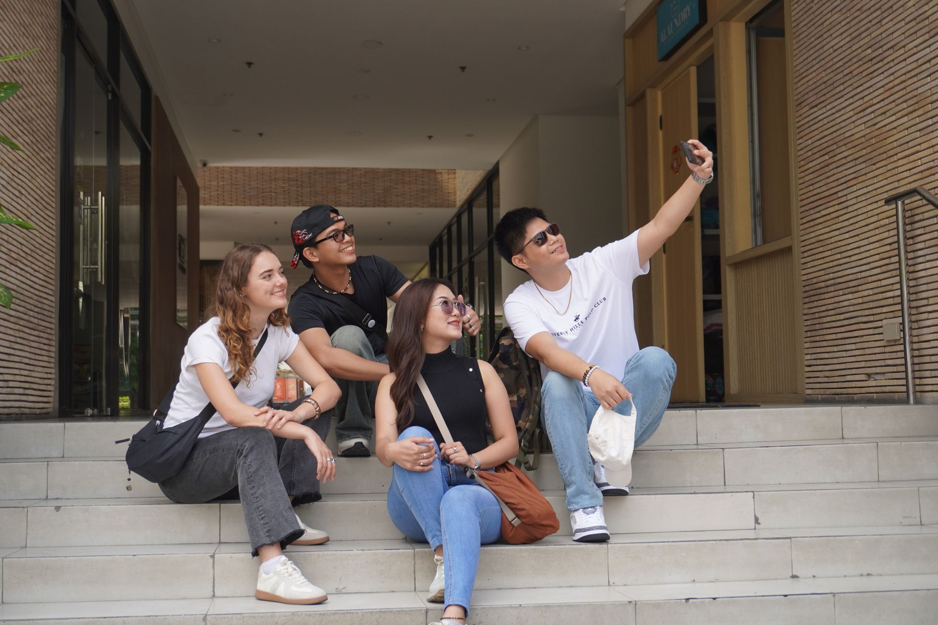 Four people taking a selfie on steps outside a building; one person holds the phone.