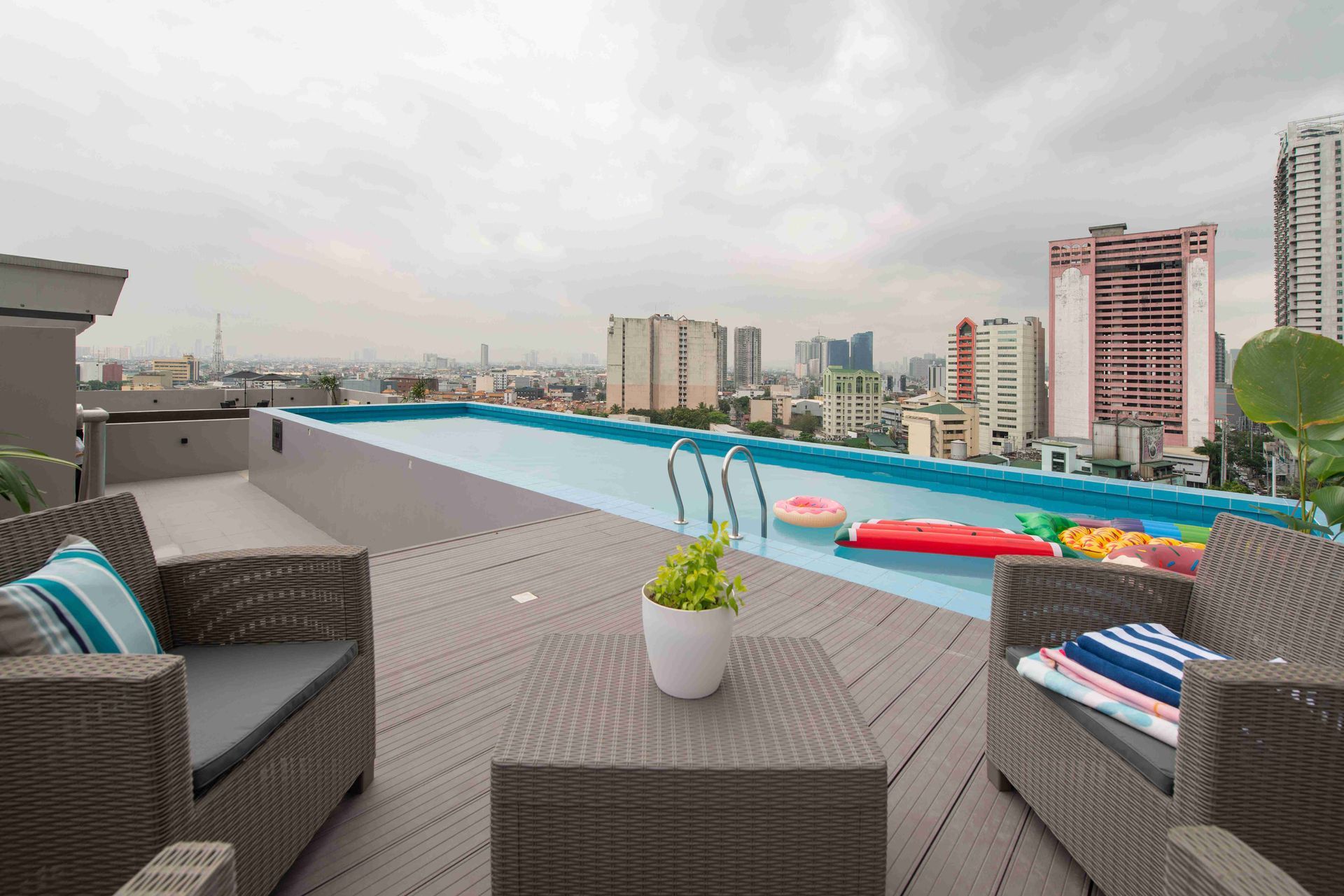Rooftop pool with city view; wicker furniture, blue cushions, and colorful floaties.