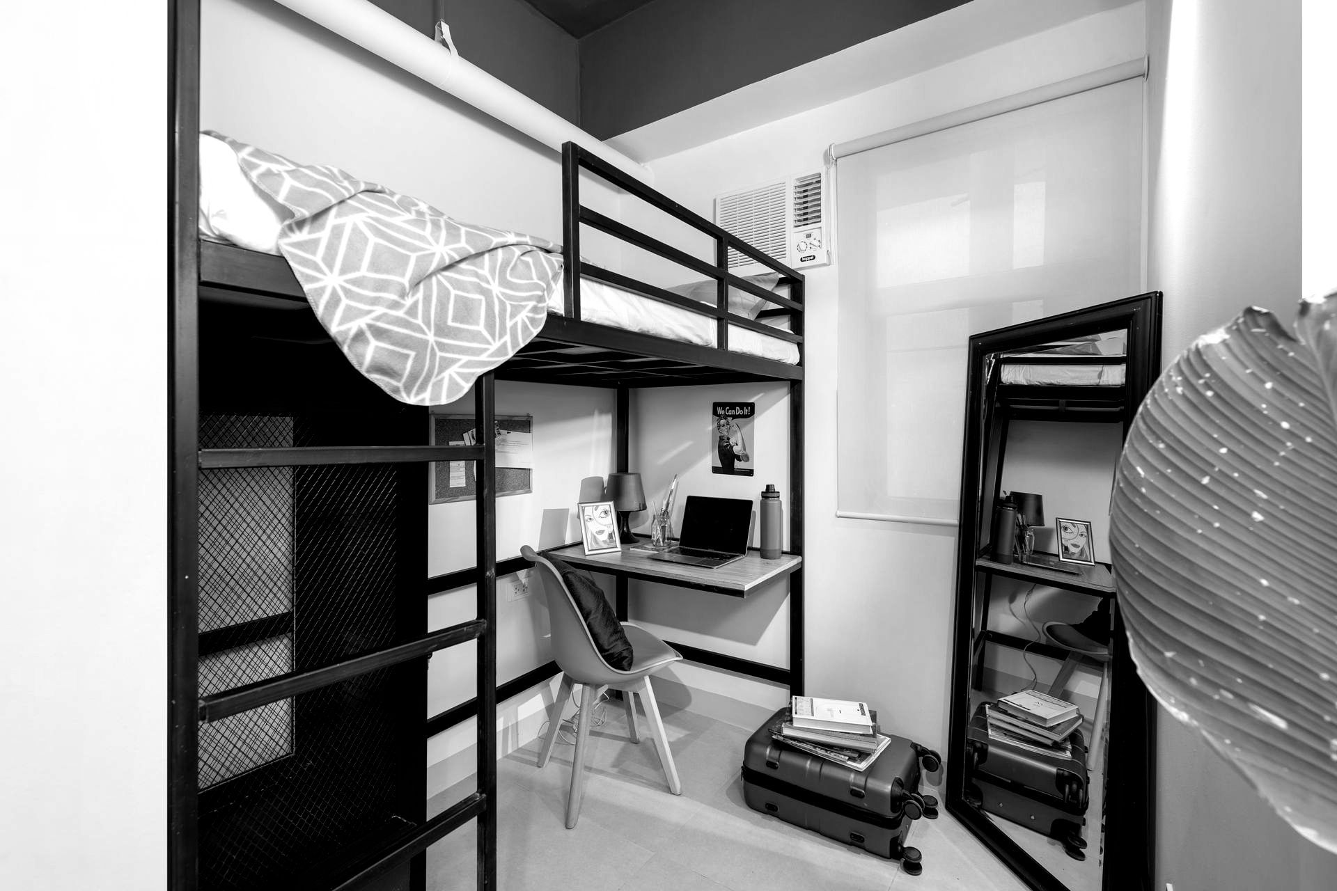 Black and white dorm room with bunk bed, desk, chair, shelves, luggage, and window.