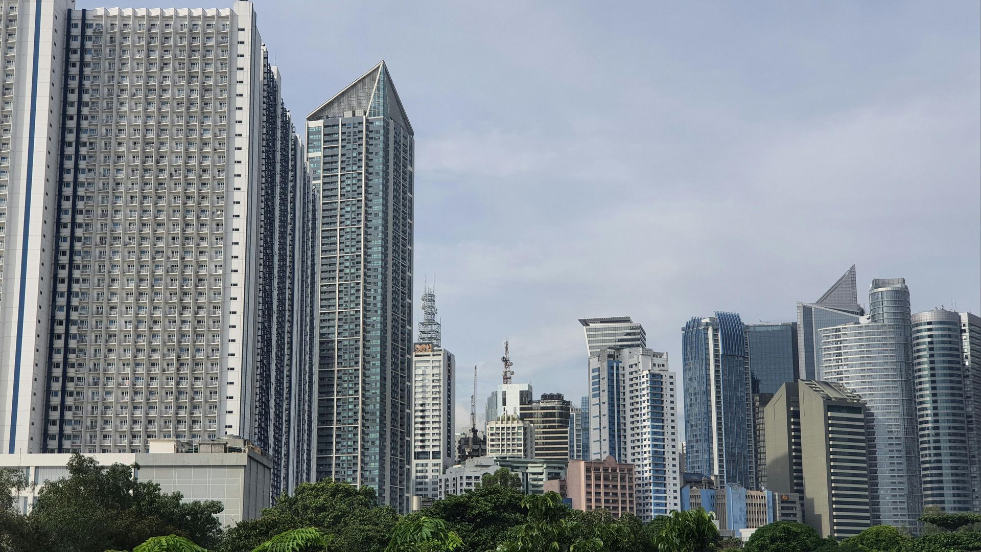 City skyline with modern high-rise buildings against a cloudy sky. Trees in the foreground.