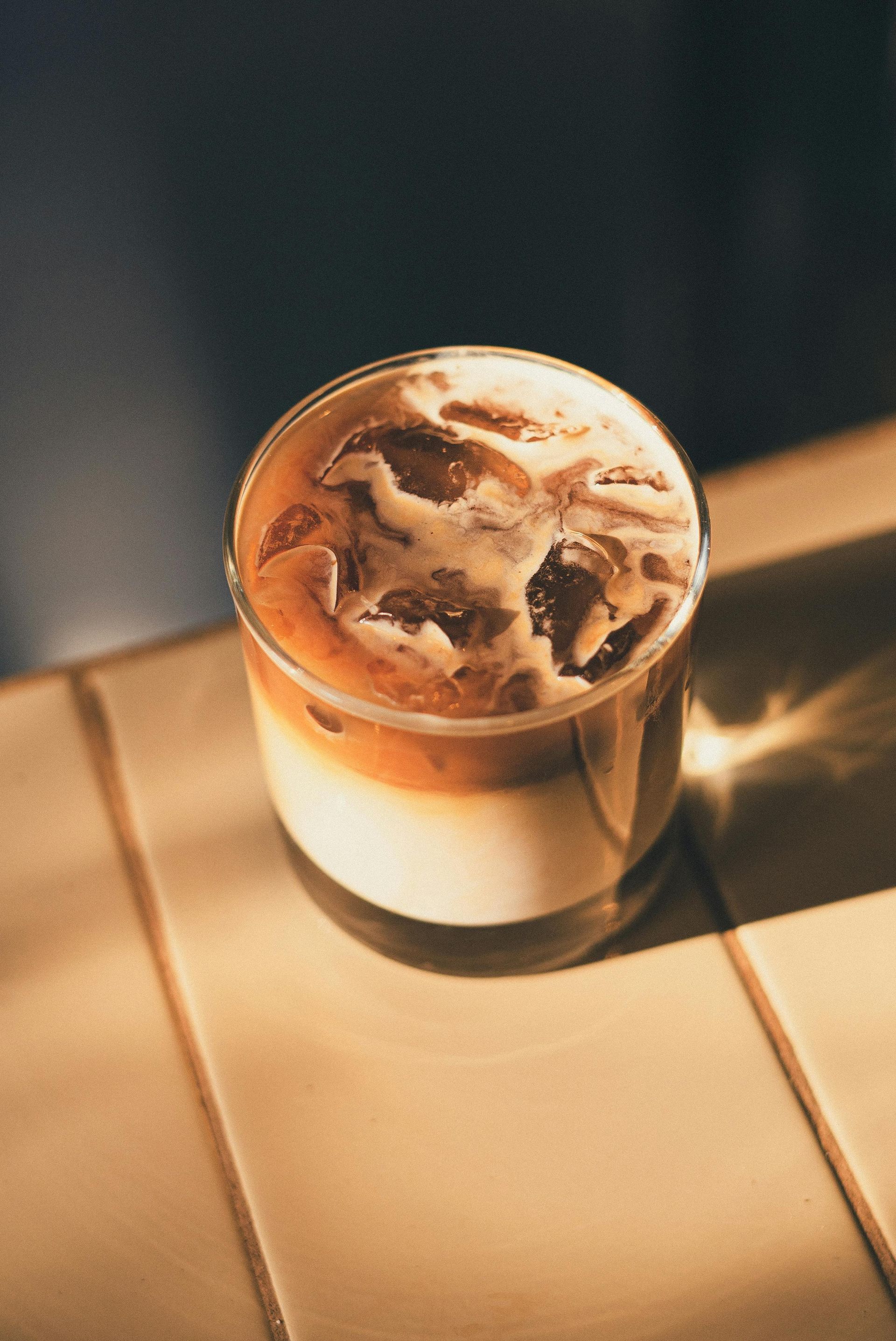 An iced latte in a glass tumbler sits on a tiled surface, illuminated by warm, angled sunlight.