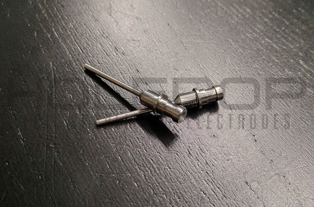Small silver metal tool or connector on a dark textured surface, with two thin prongs extending left.