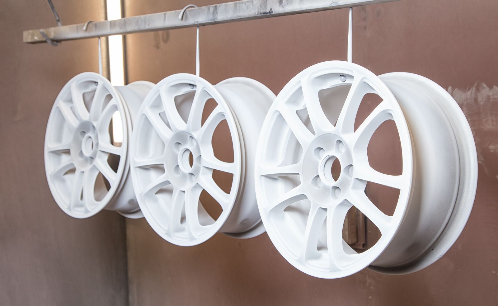 Four white car rims hanging from a rack in a spray-painting booth.