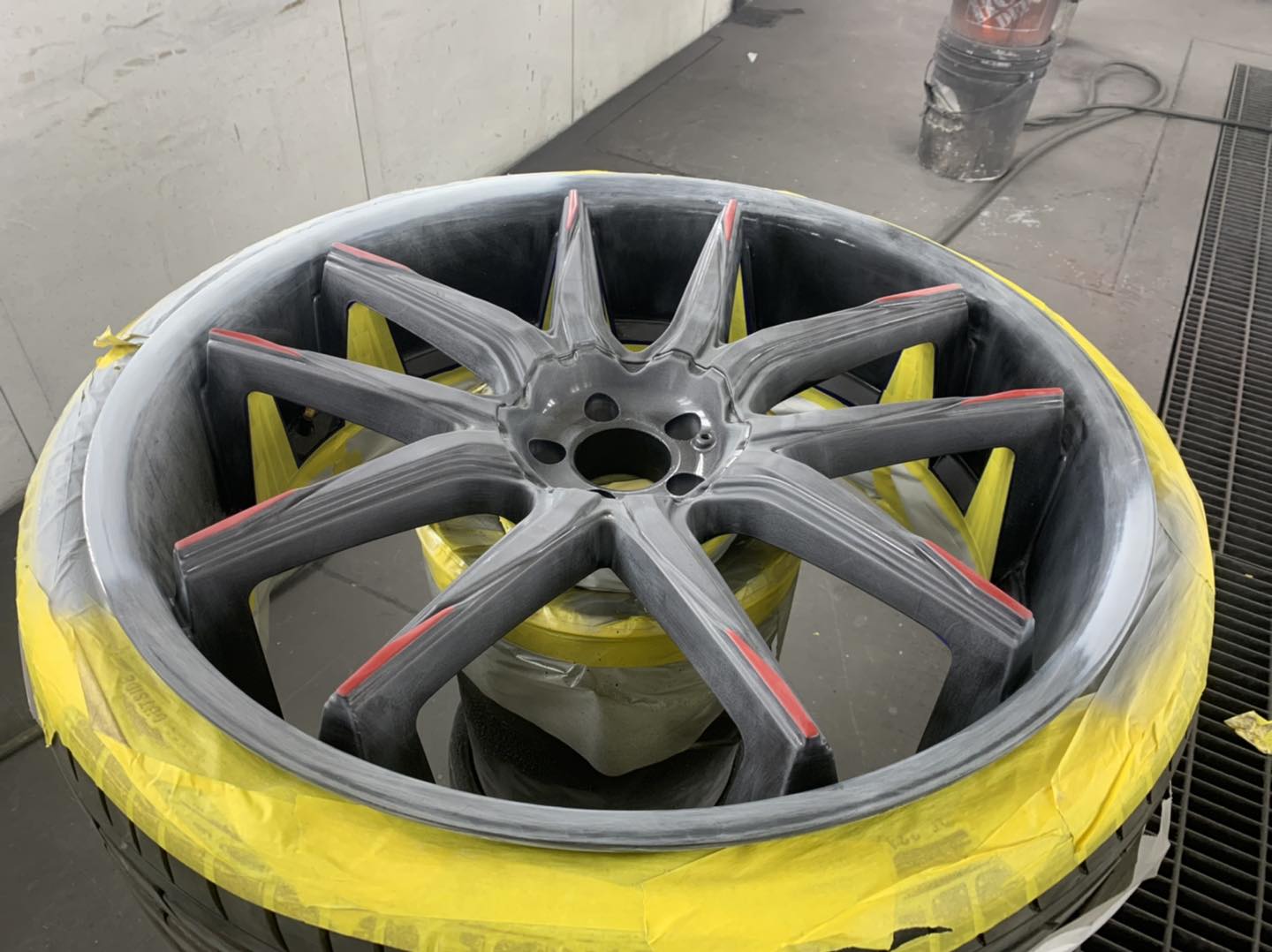 Painted car wheel, gray with red accents, yellow taped tire, in a spray booth.