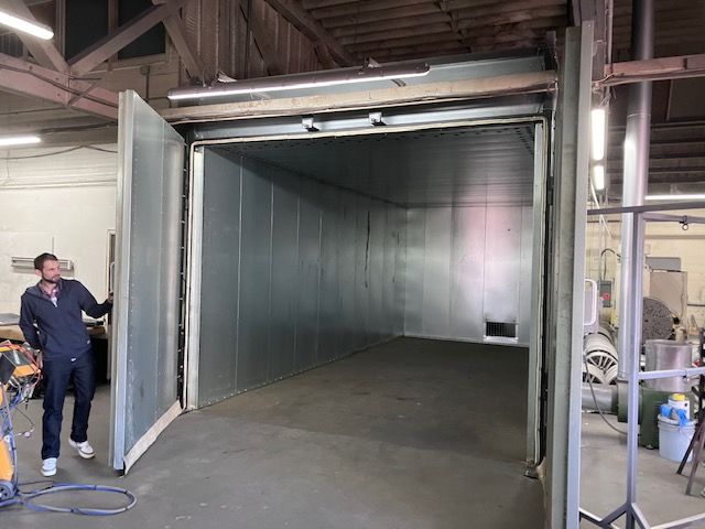 A man is standing in a room with a large furnace/oven door open used for powder coating.