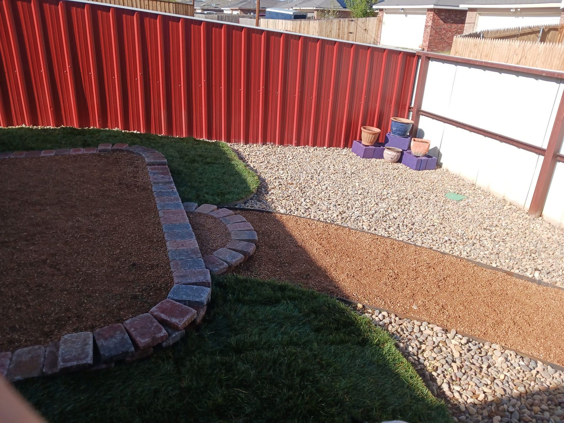 A backyard with red and white fences, grass, mulch, gravel, and decorative brick edging.