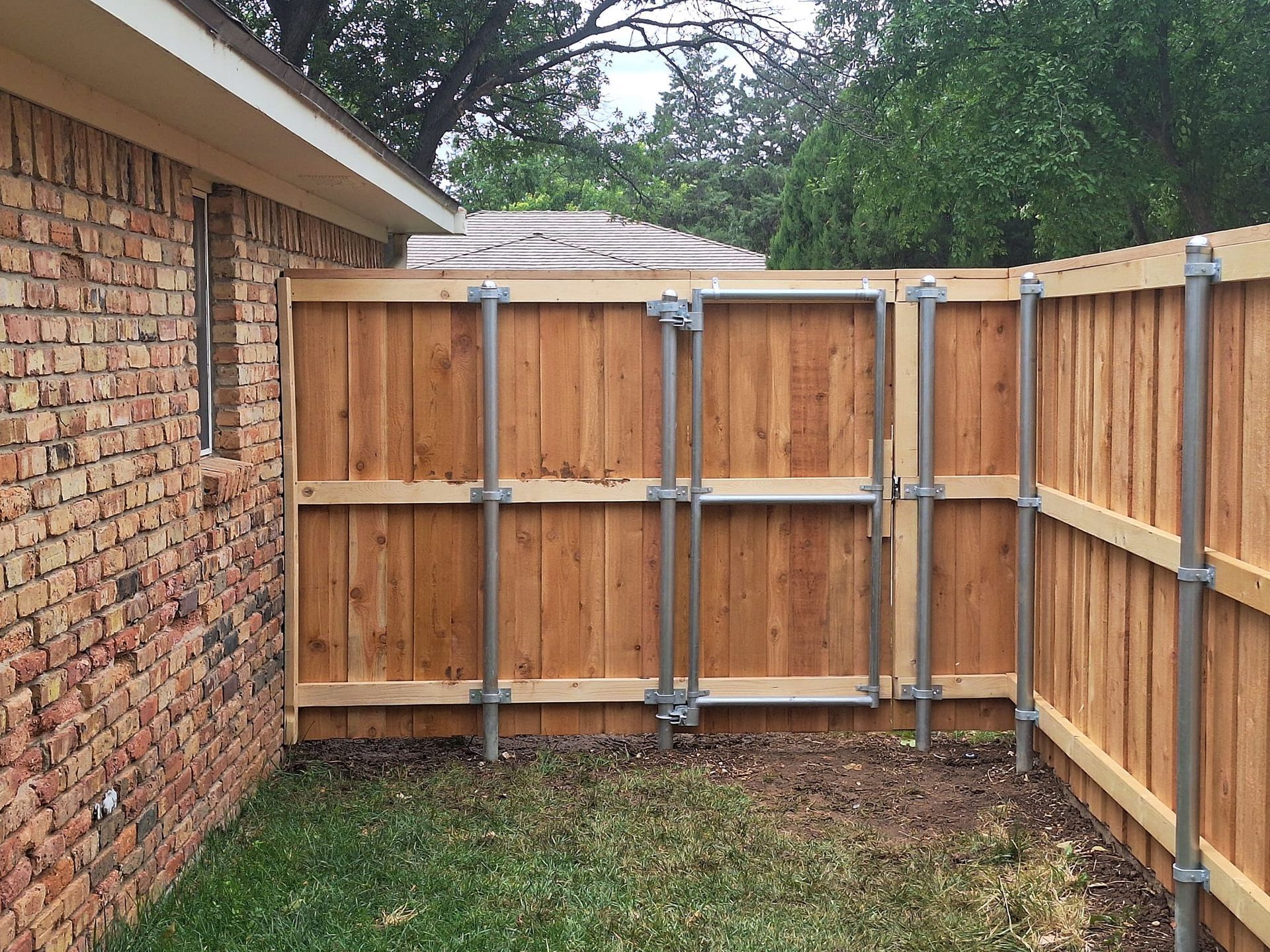 Wooden fence with metal supports installed in a grassy backyard, adjacent to a brick wall.