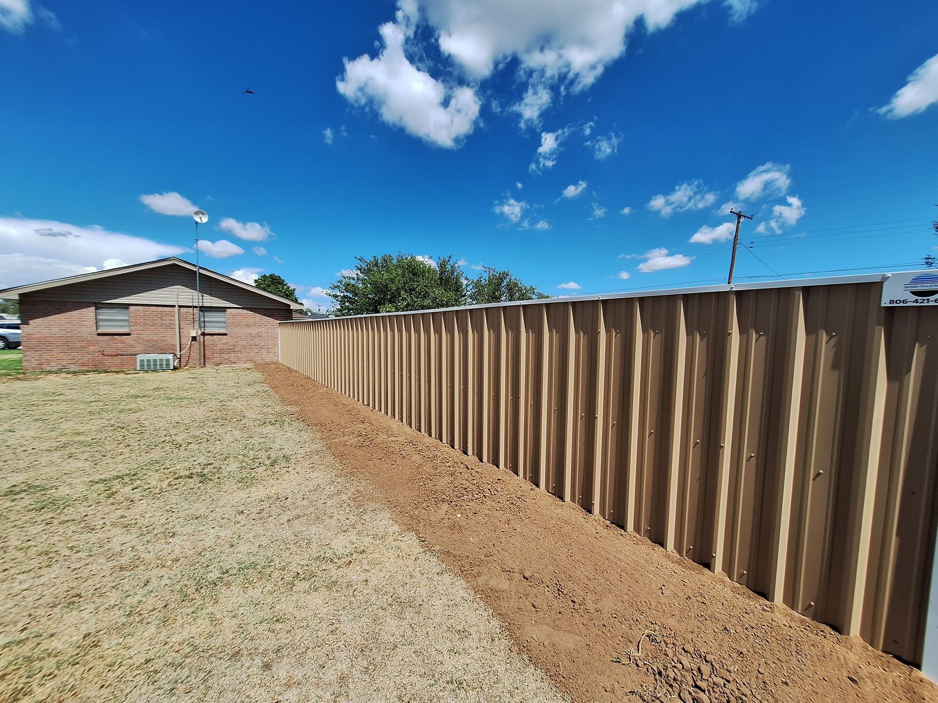 A wooden fence along a yard with a dirt trench in front, under a blue sky with clouds.