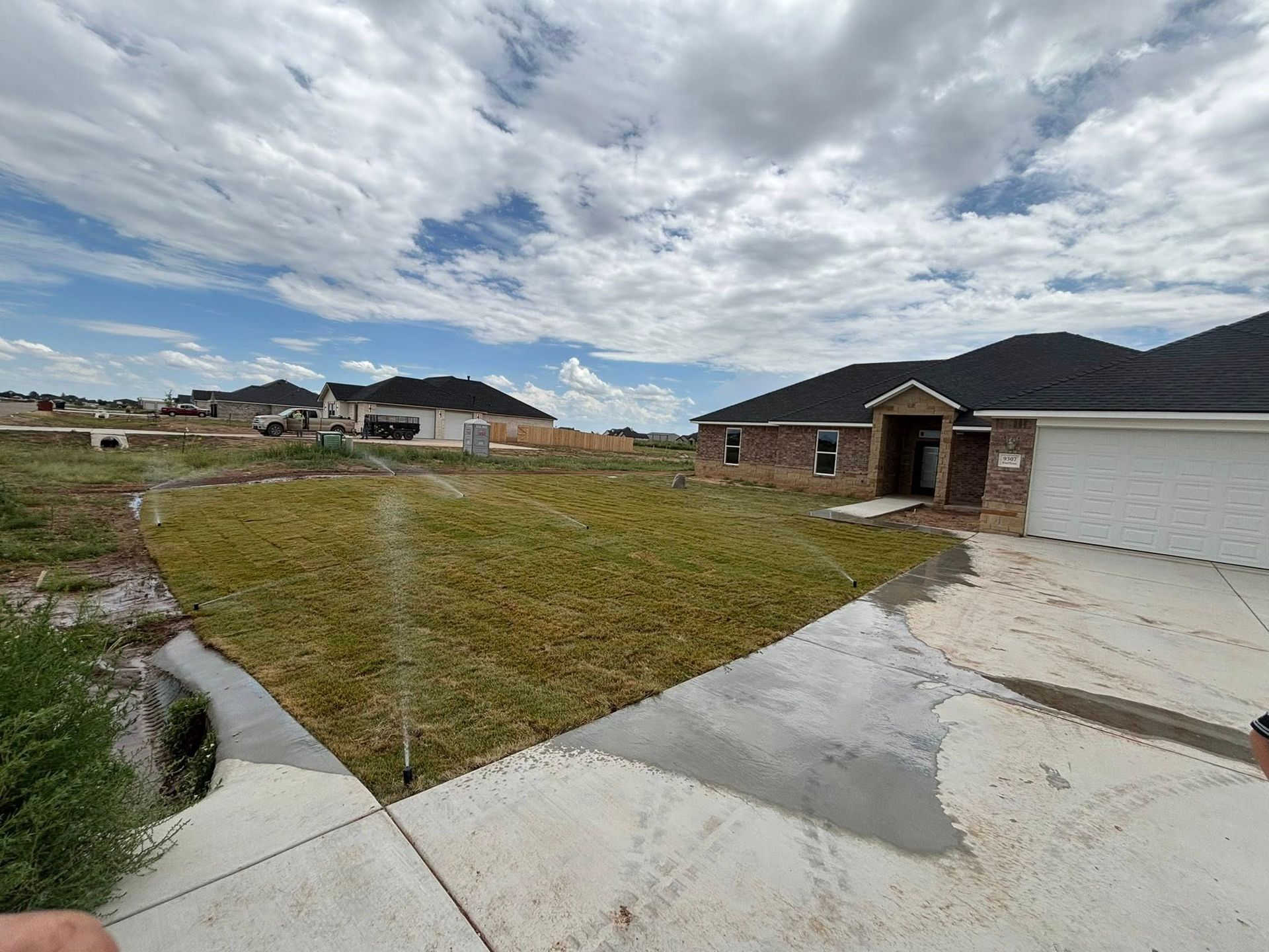 A new house with a brown brick exterior and dark roof; lawn sprinklers watering the grassy yard under a cloudy sky.