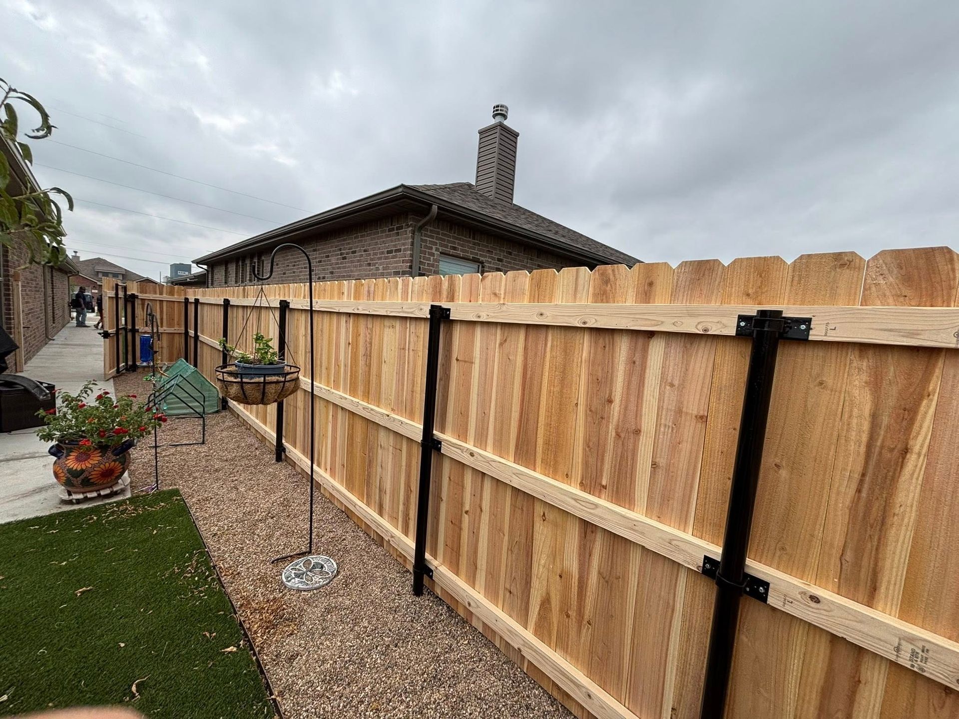 Wooden fence with black metal posts, gravel bed, and potted plants against a brick house on a cloudy day.