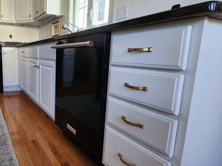 Black dishwasher beside white kitchen cabinets with gray drawers and brass pulls on a wood floor