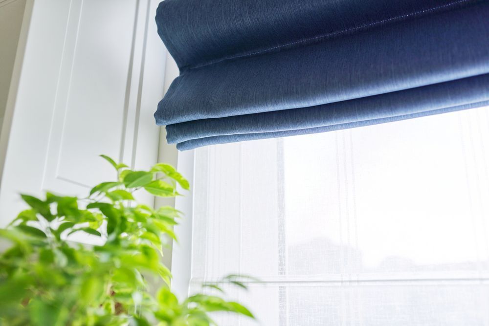 A Blue Roman Shade is Hanging on a Window Next to a Plant — Curtain Haven & Blinds in Laurieton, NSW